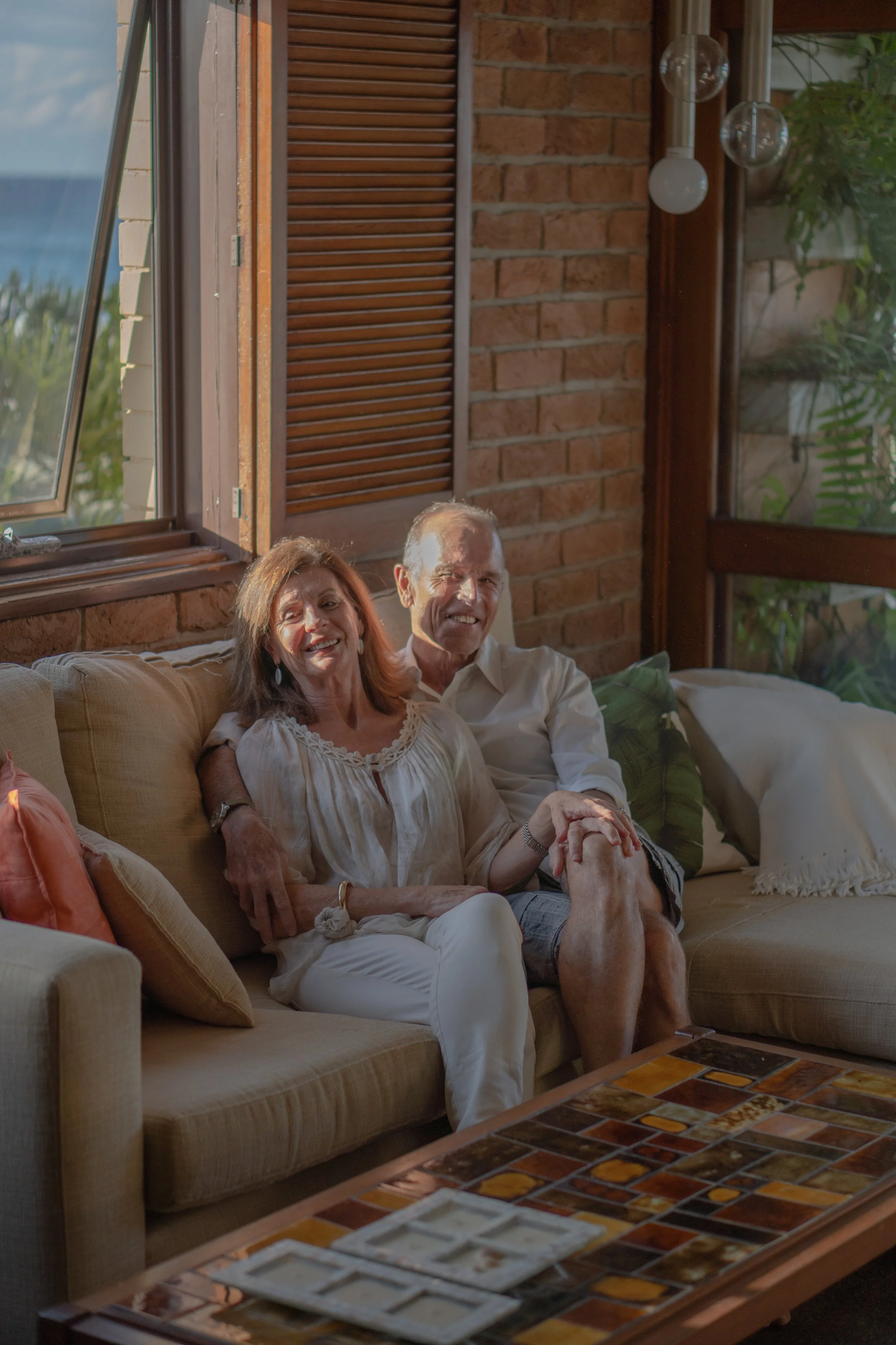 An elderly couple sitting on a beige sofa inside a room with brick walls and large windows, smiling and holding hands.