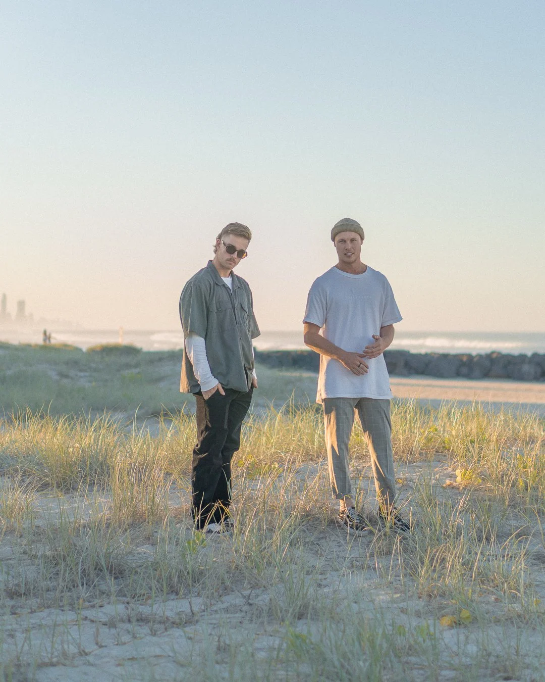 Two men standing on a grassy beach during sunset, with ocean and city skyline in the background.