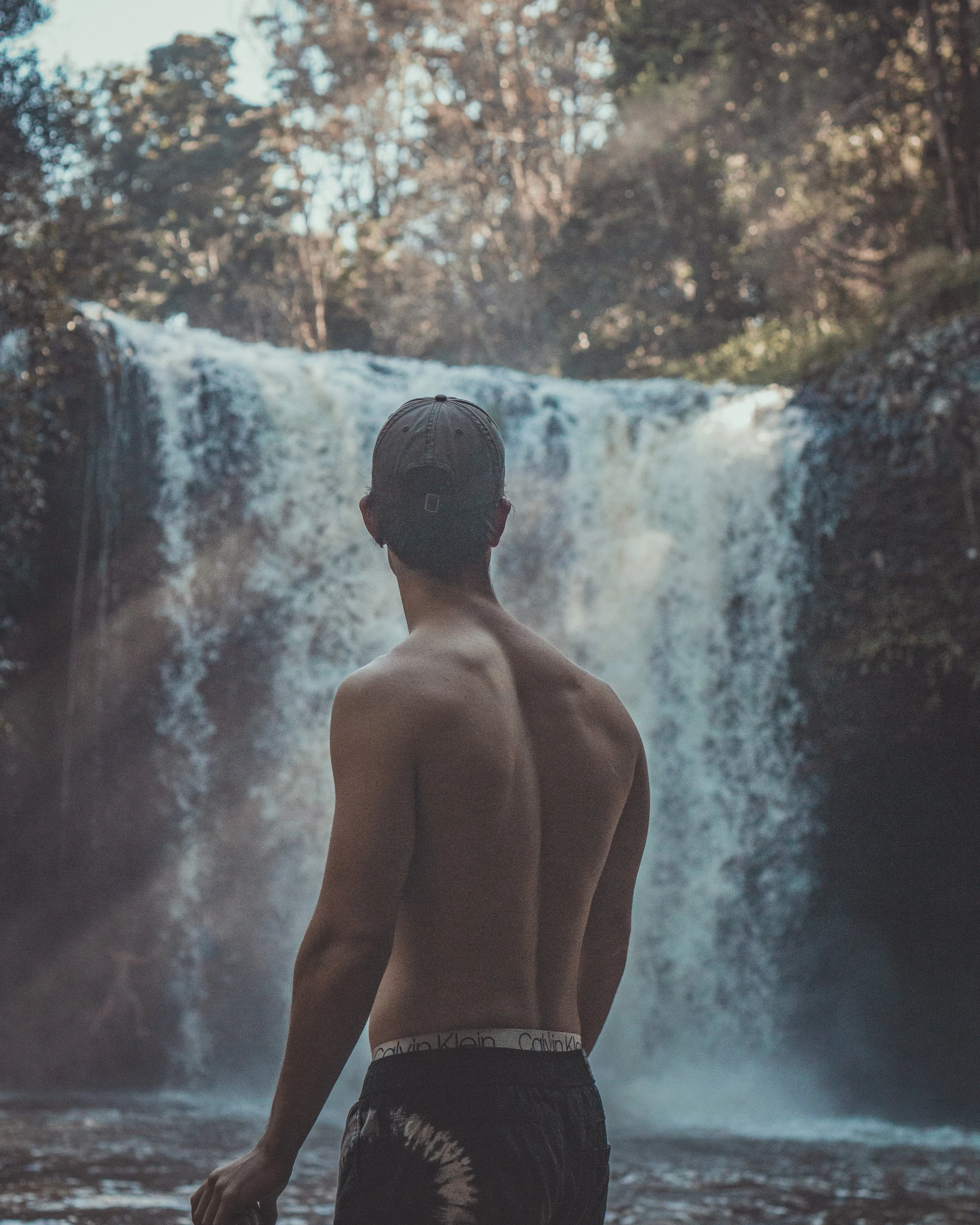 A shirtless man wearing a backwards cap stands near a waterfall, with trees and rocks in the background.