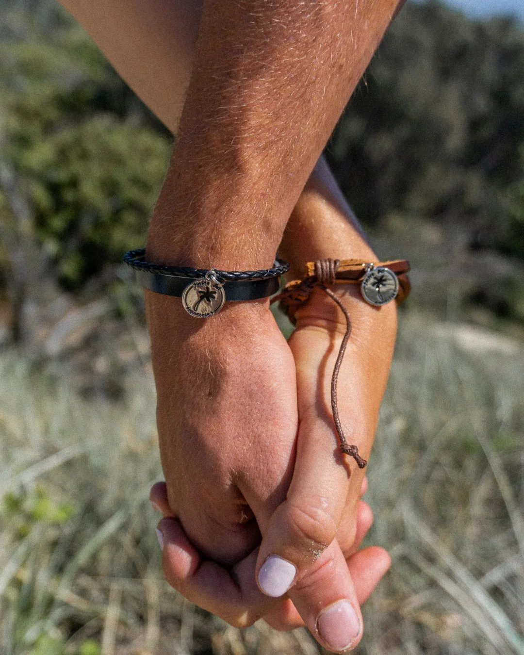 Two people holding hands, each wearing a bracelet with a palm tree charm, outdoors with blurry greenery in the background.