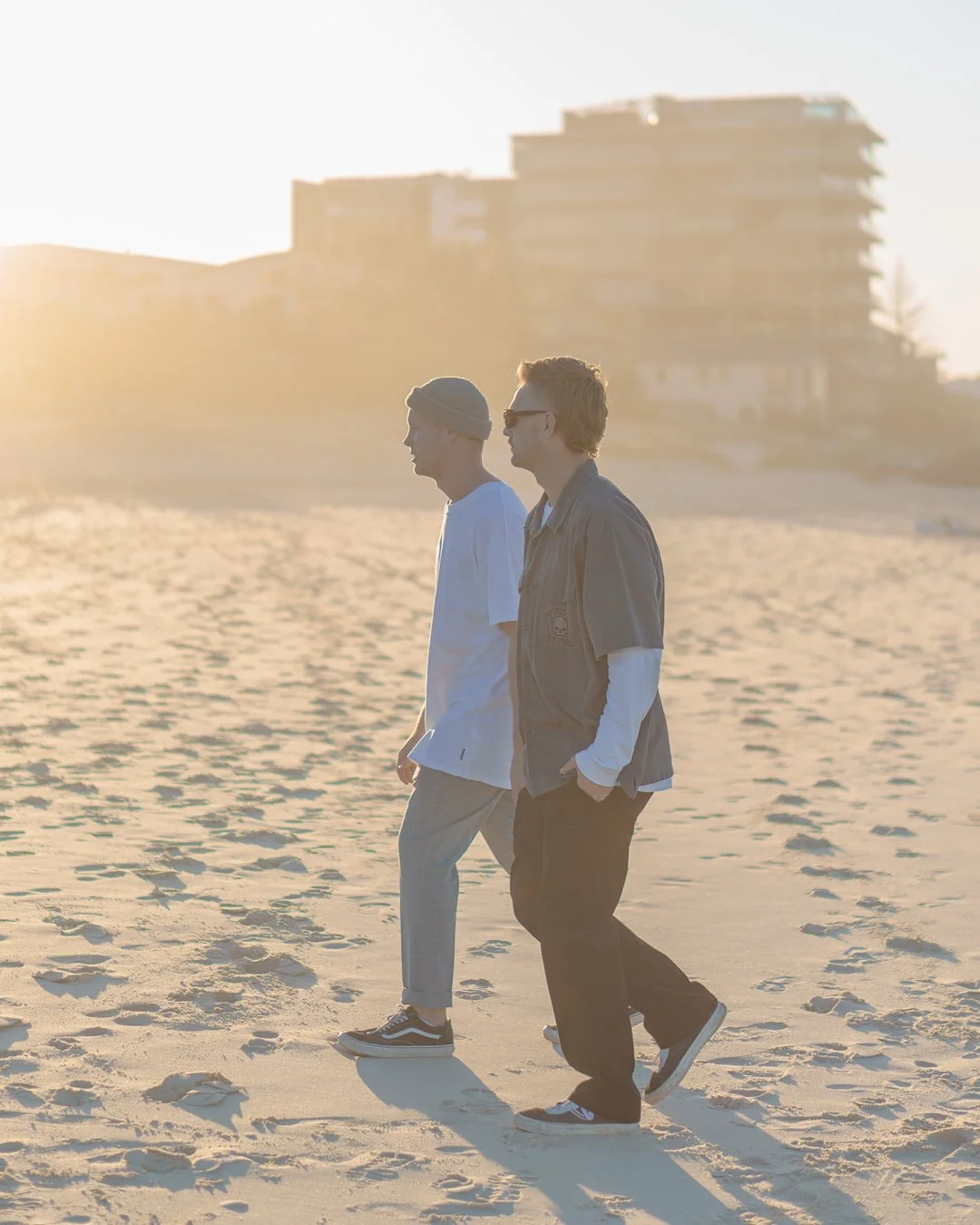 Two young men walking on a sandy beach during sunset, with buildings in the background. At Gold Coast
