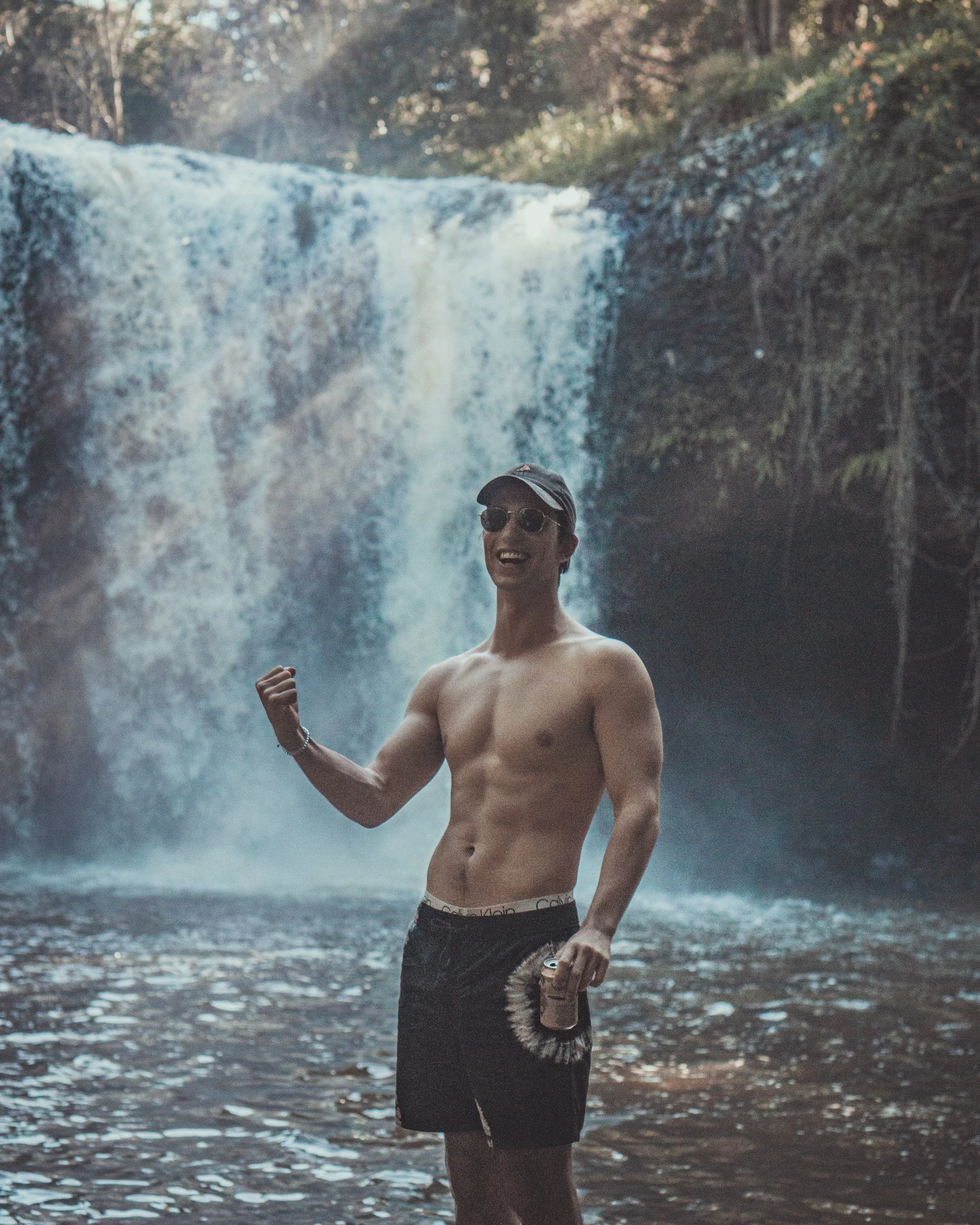A shirtless man wearing sunglasses, a cap, and shorts, smiling and flexing his arm while holding a drink, standing in front of a waterfall.