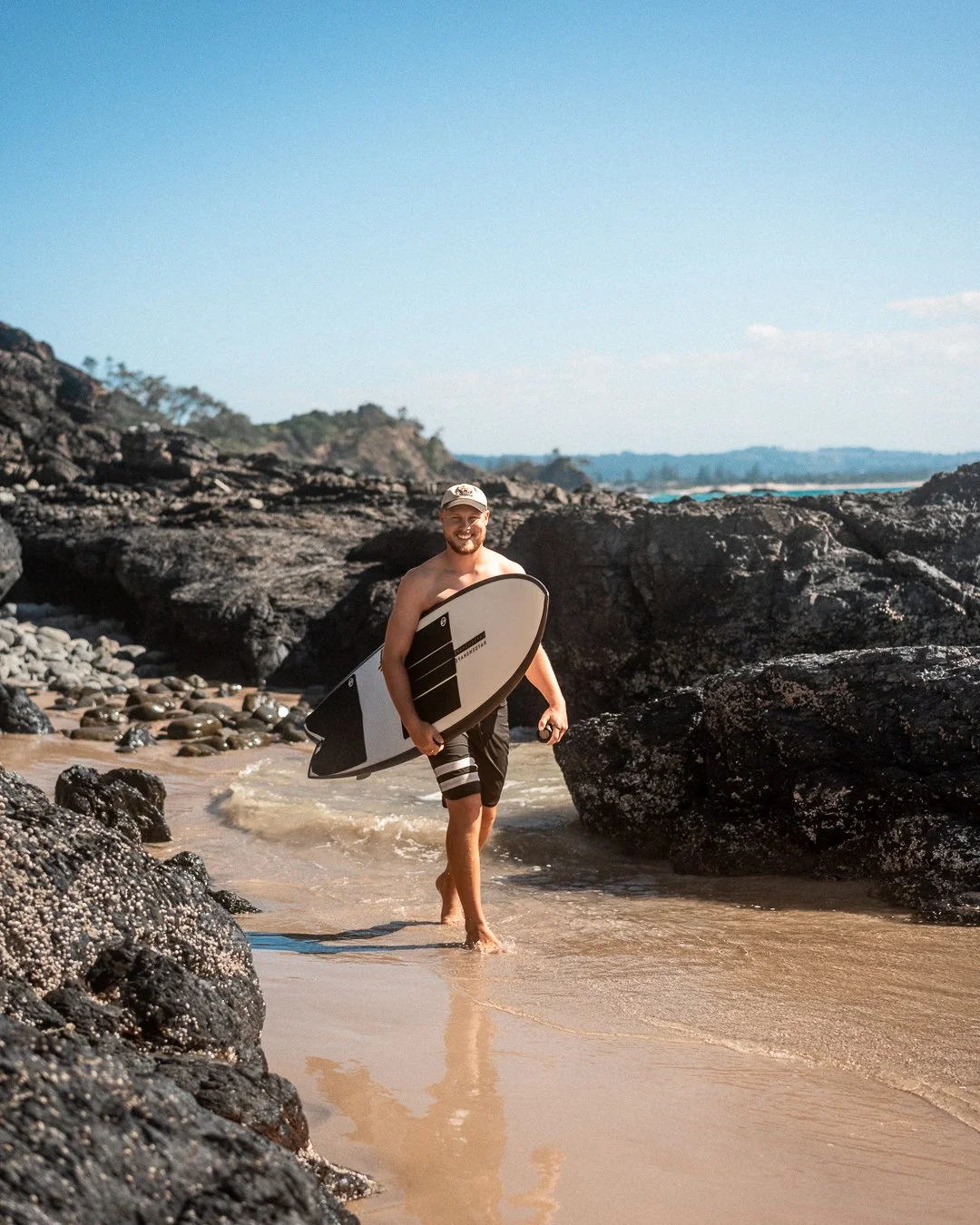 A man carrying a surfboard walking out of the water onto a rocky beach with sand and large rocks around him, under a clear blue sky.