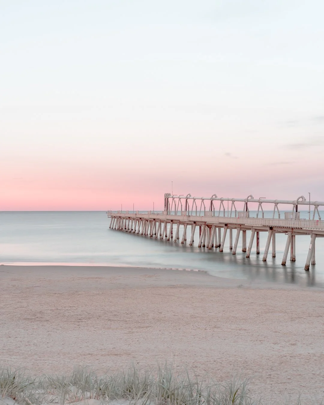 A pier at Main Beach on the Gold Coast extending into the ocean at sunset with pastel pink and blue sky, calm waters, sandy beach, and some grass in the foreground.