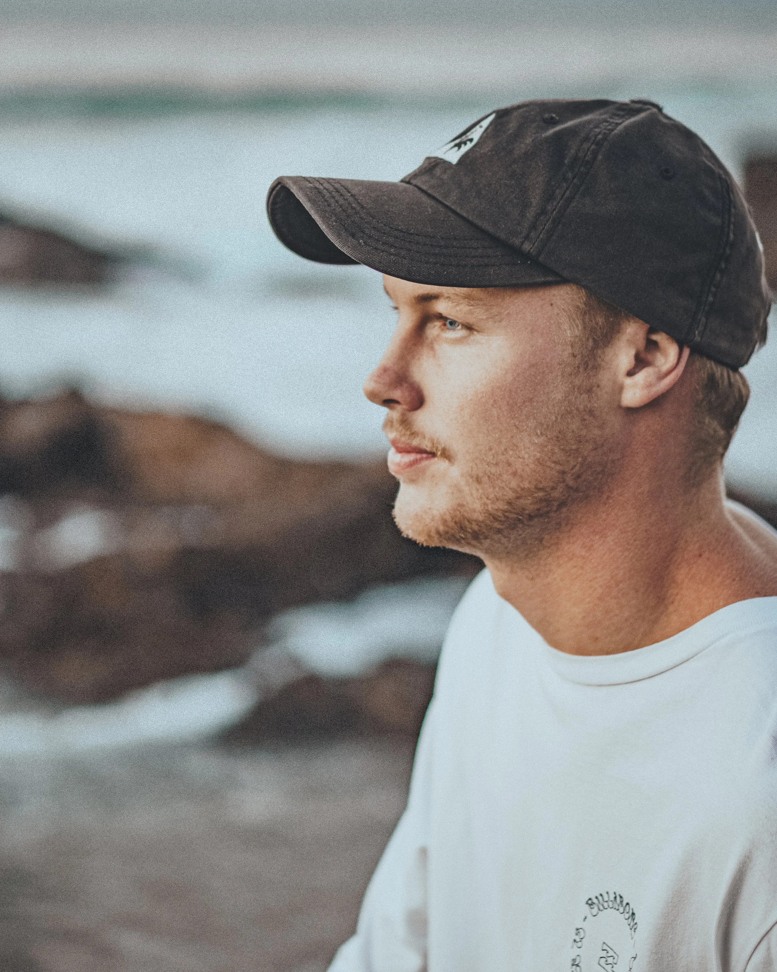A young man with blond hair and light facial hair wearing a black baseball cap and white t-shirt, outdoors near water.