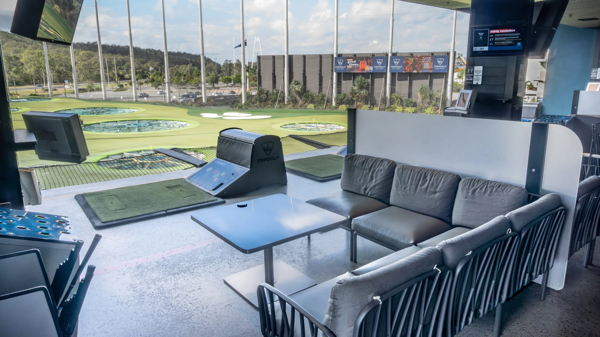 Indoor golf simulation room with a view of outdoor mini golf course through large windows, featuring seating area, tables, and golf simulators.