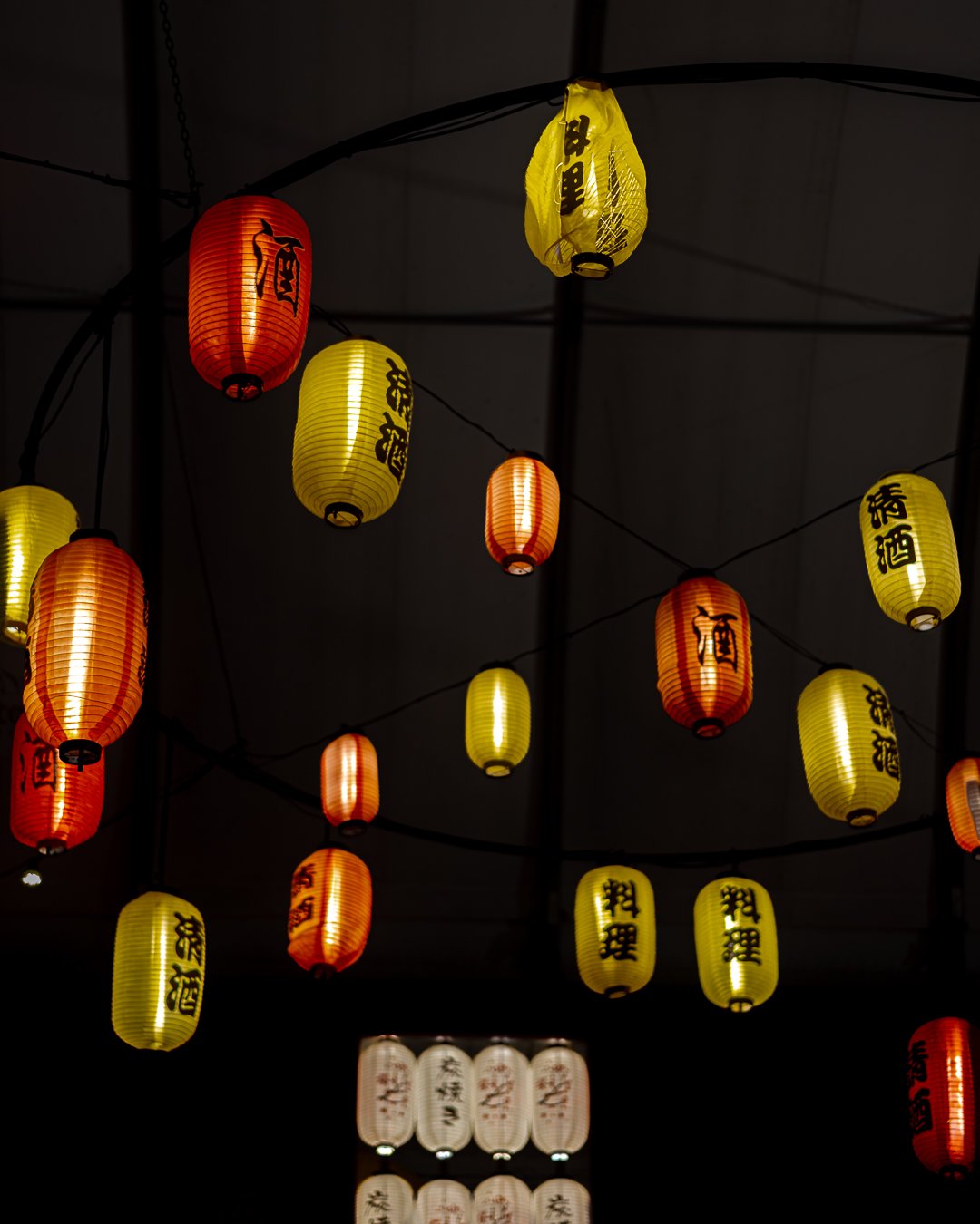 Colorful paper lanterns hanging from the ceiling at night, illuminated. The lanterns are in red, yellow, and white, with black Asian characters written on them.