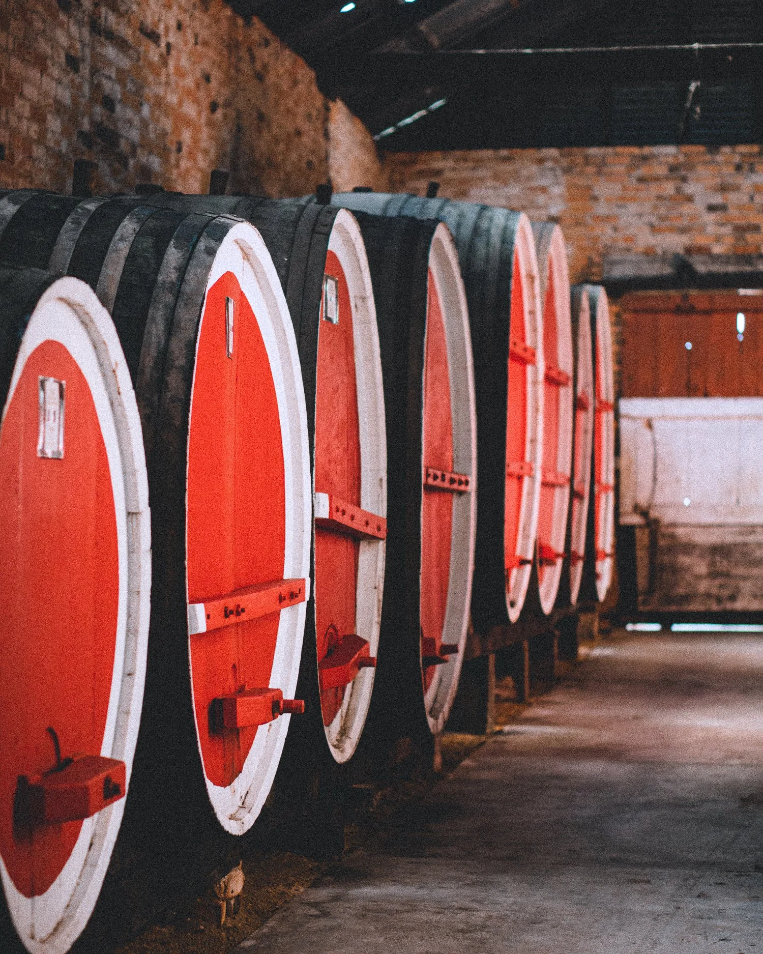 Rows of wine barrels with red tops stored in a rustic cellar with exposed brick walls.
