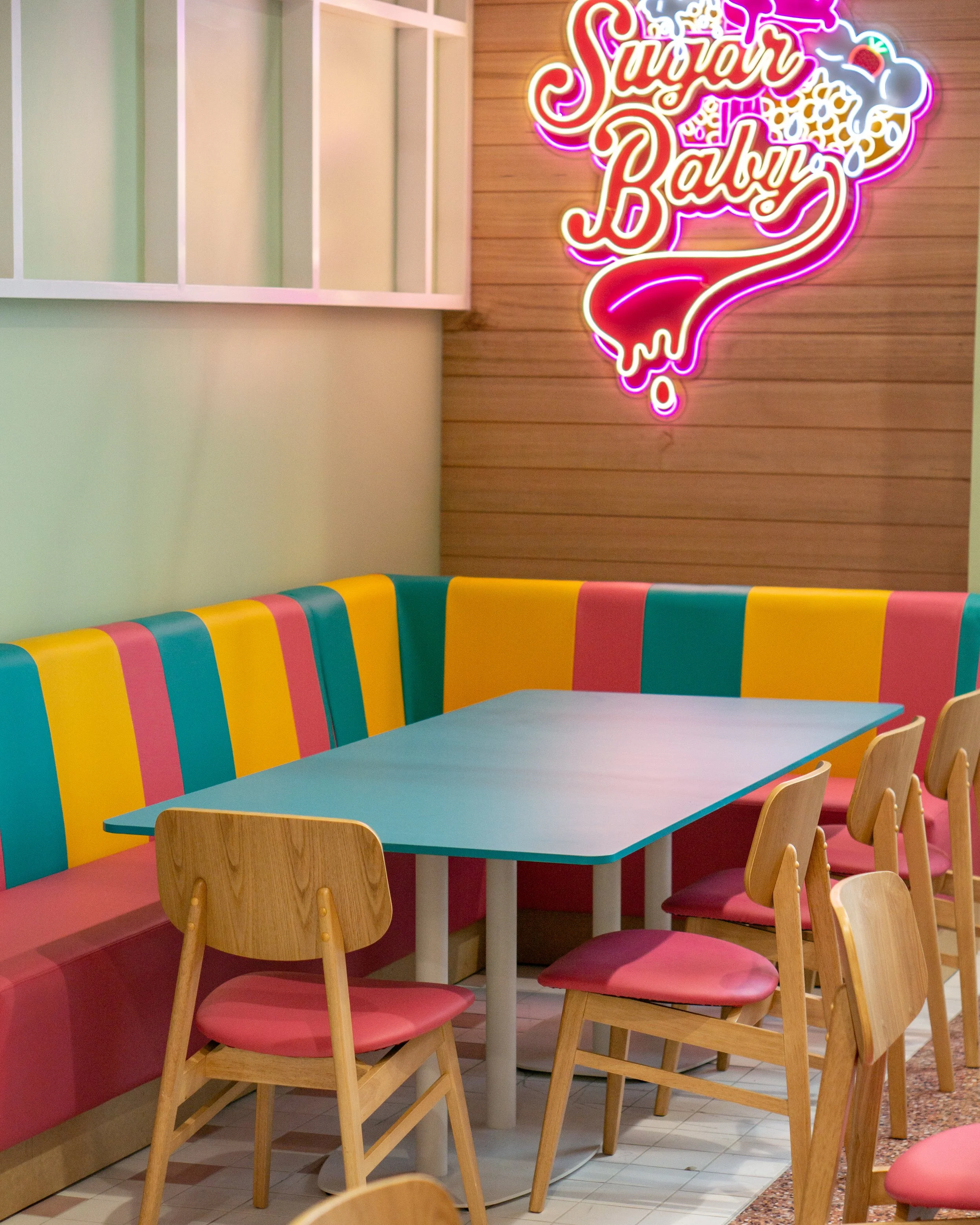 Colorful pastel decor in a sweet-themed cafe with a neon sign reading 'Sugar Baby', a pastel blue table, pink and yellow seating, and wooden chairs.