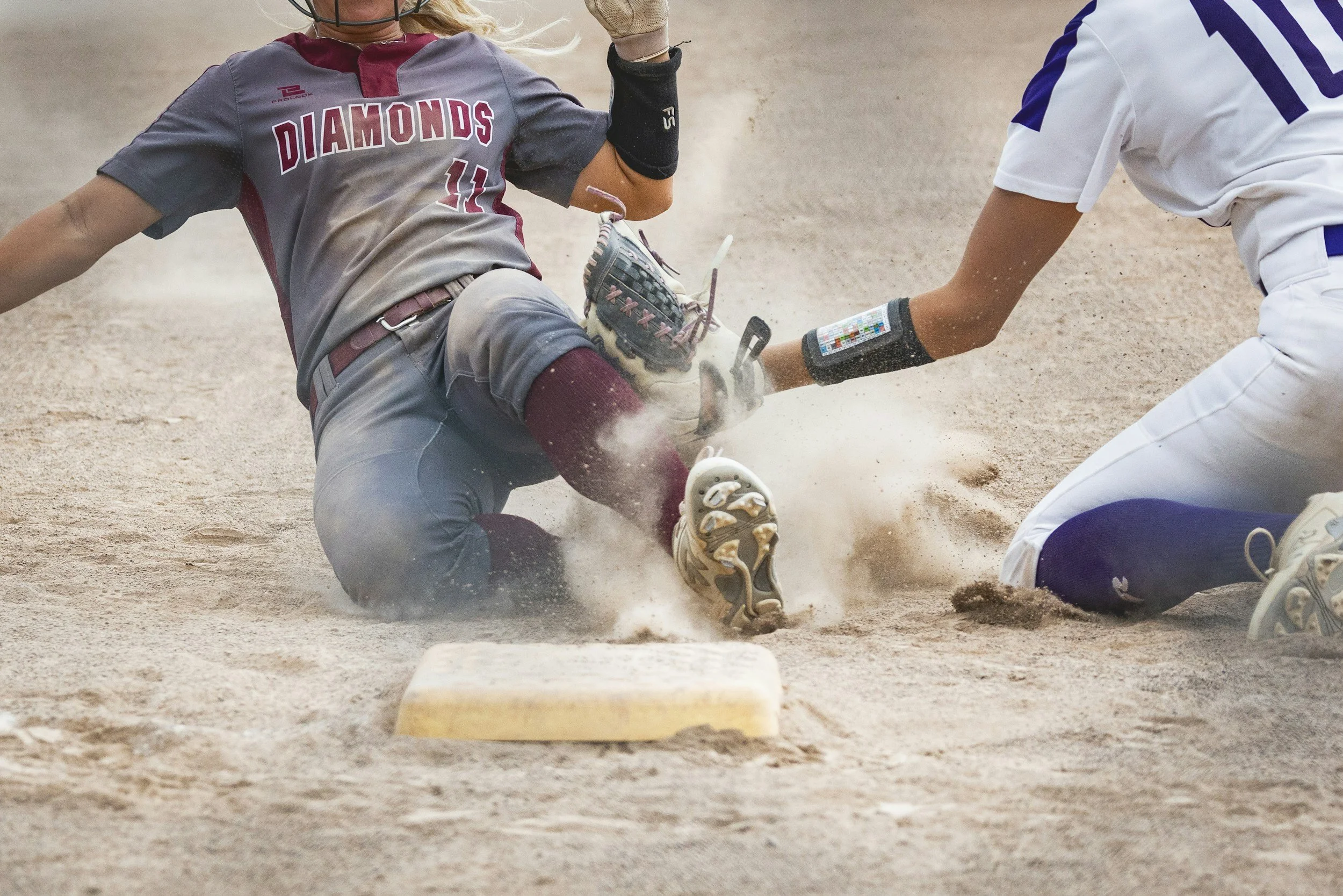A baseball player wearing a grey and maroon uniform with the number 11, sliding headfirst into a base while an opposing player in a white and blue uniform attempts to tag him out. Dust is flying from the dirt around the players.