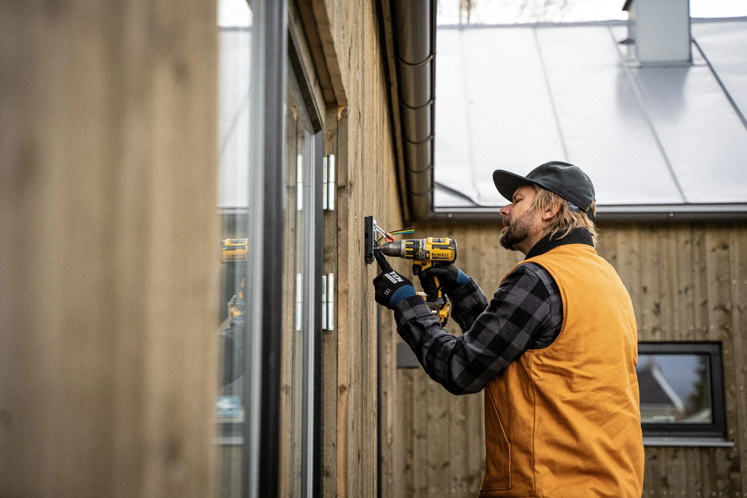 A handyman wearing a black cap, black gloves, a brown vest, and a black and gray checkered shirt uses a yellow power drill to secure a window frame to an exterior wooden wall of a house.