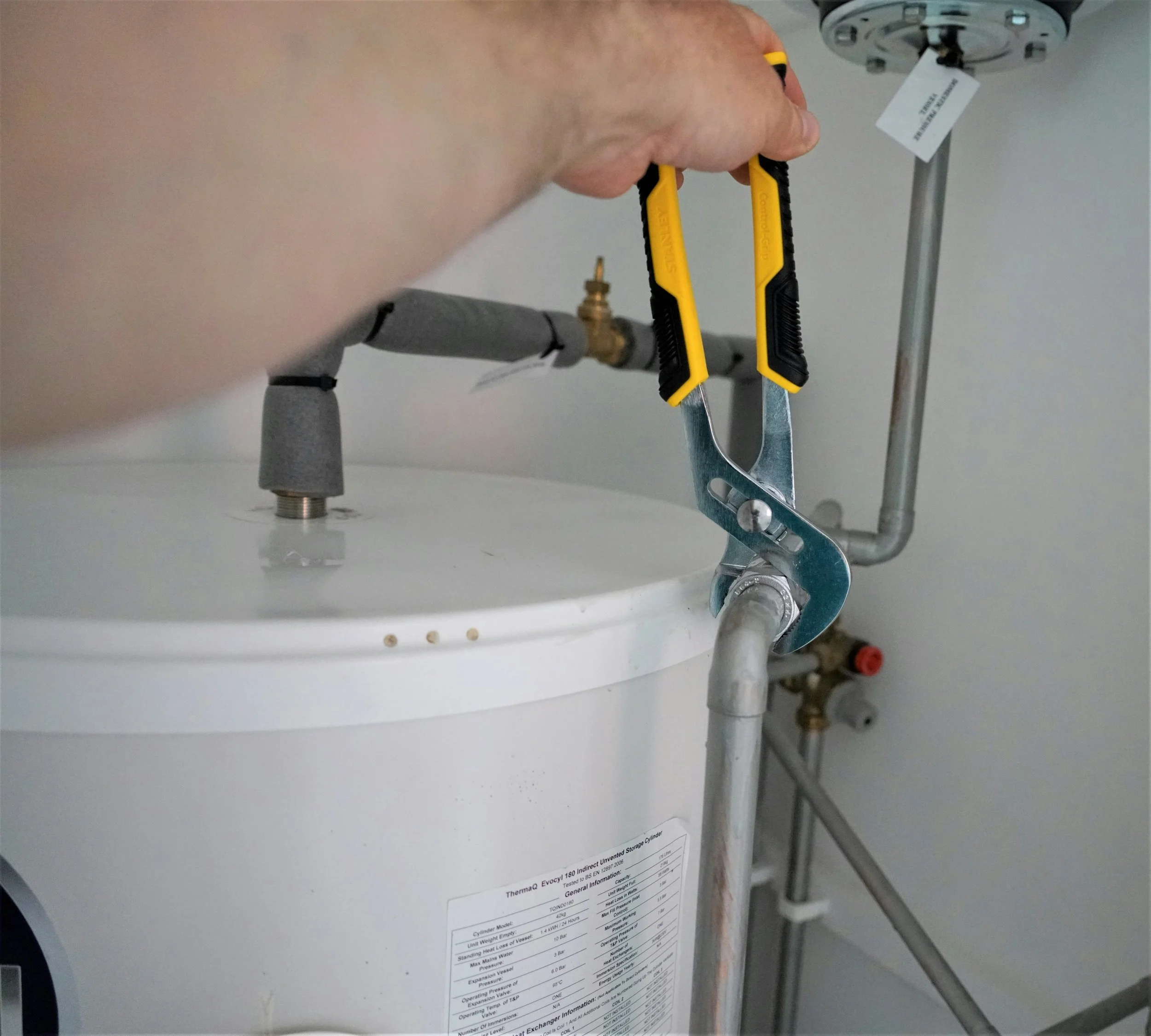 A plumber is using pliers to unscrew a pipe on a water heater in a utility room.