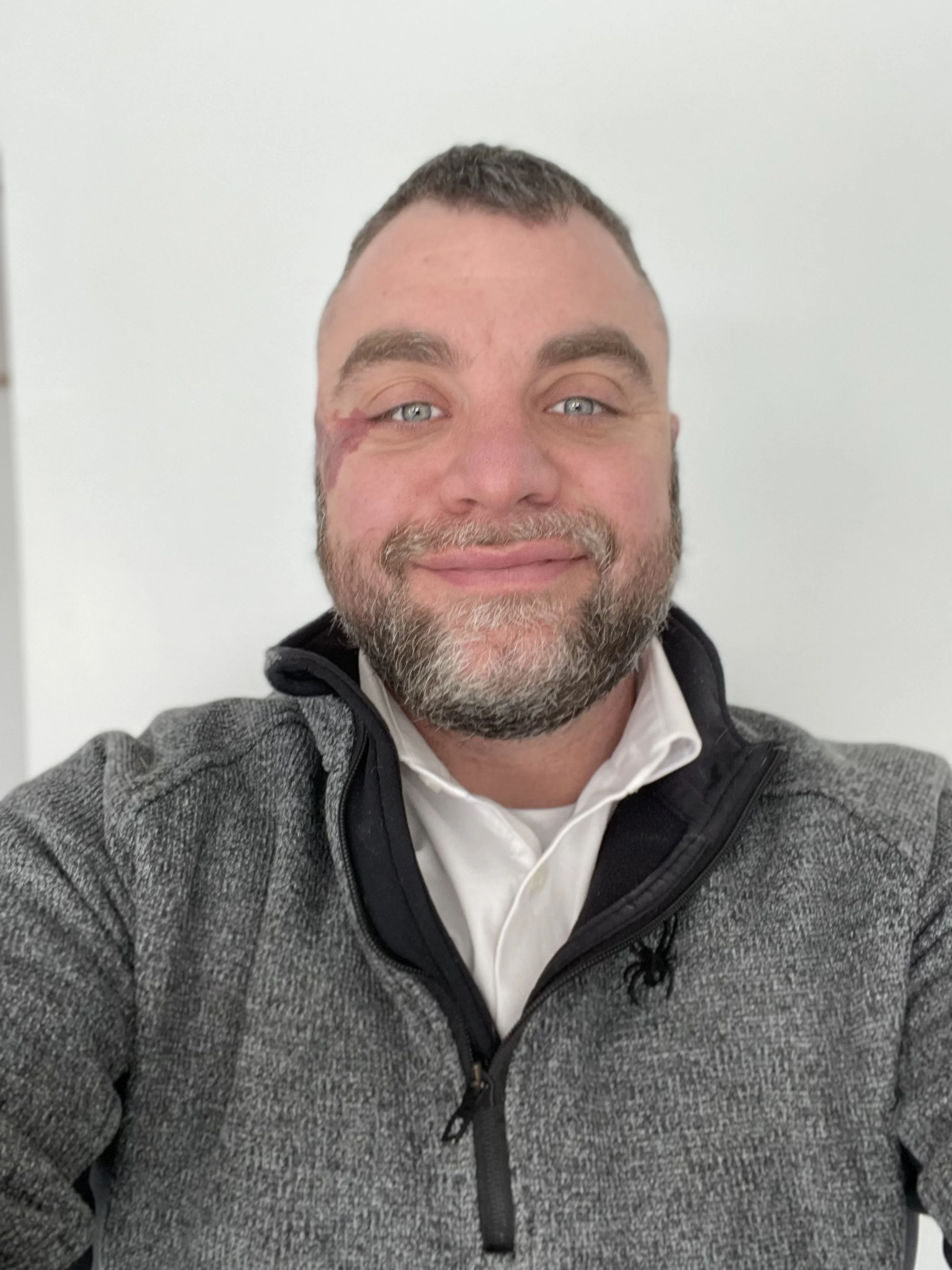 A man with a beard, gray hair, and light blue eyes taking a selfie in front of a plain white wall. He is wearing a white shirt and a gray zip-up jacket.
