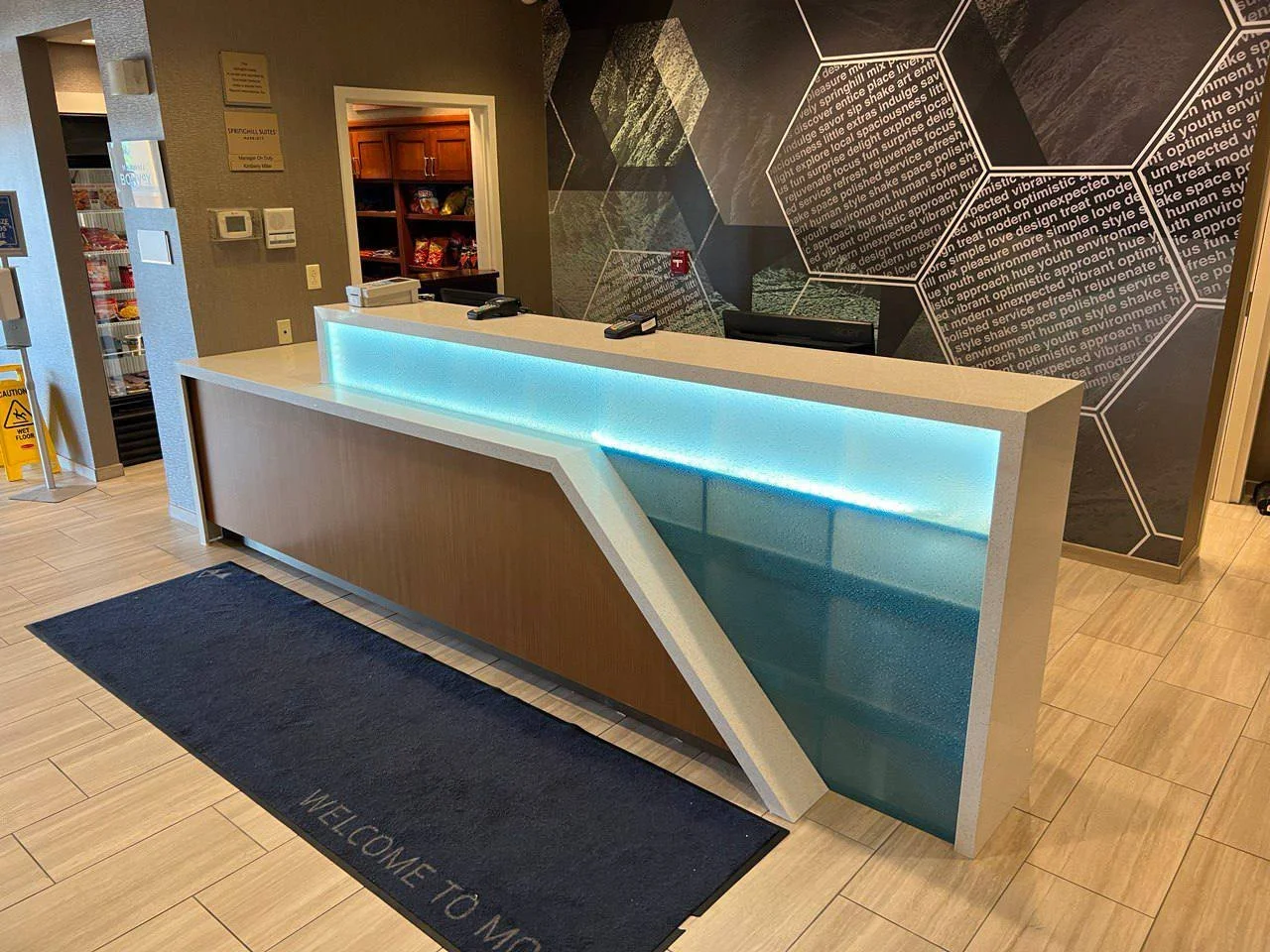 Hotel reception desk with modern design and illuminated counter, hexagonal wall art, and adjacent snack area.