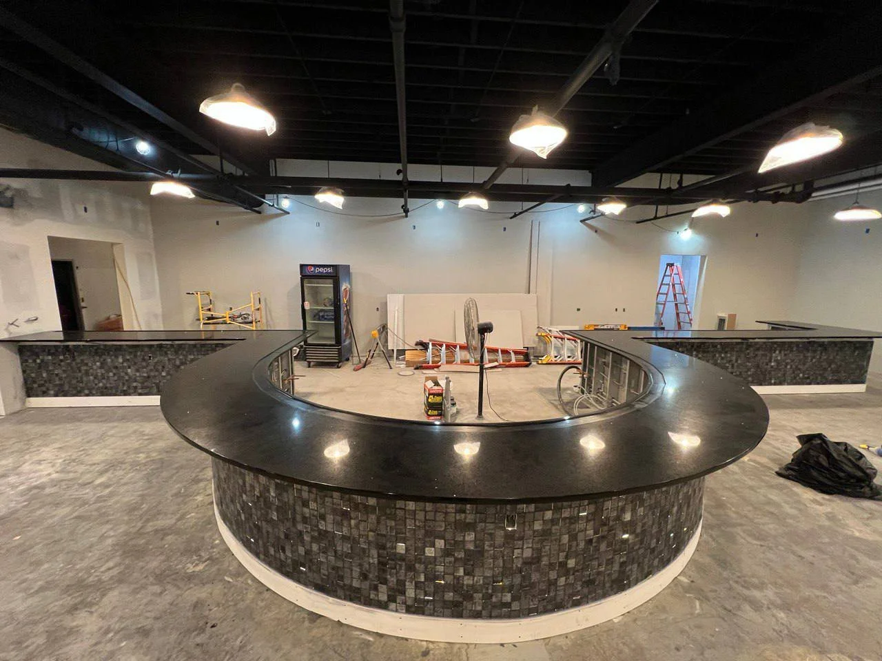 Interior of a partially constructed bar area with a large curved countertop, pendant lights above, and a tiled bar front.