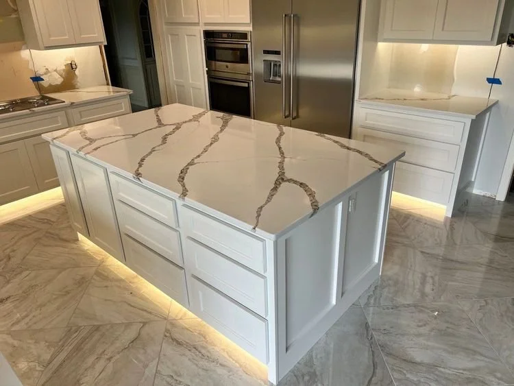 Modern kitchen with a large white marble island, stainless steel appliances, white cabinetry, and under-cabinet lighting.
