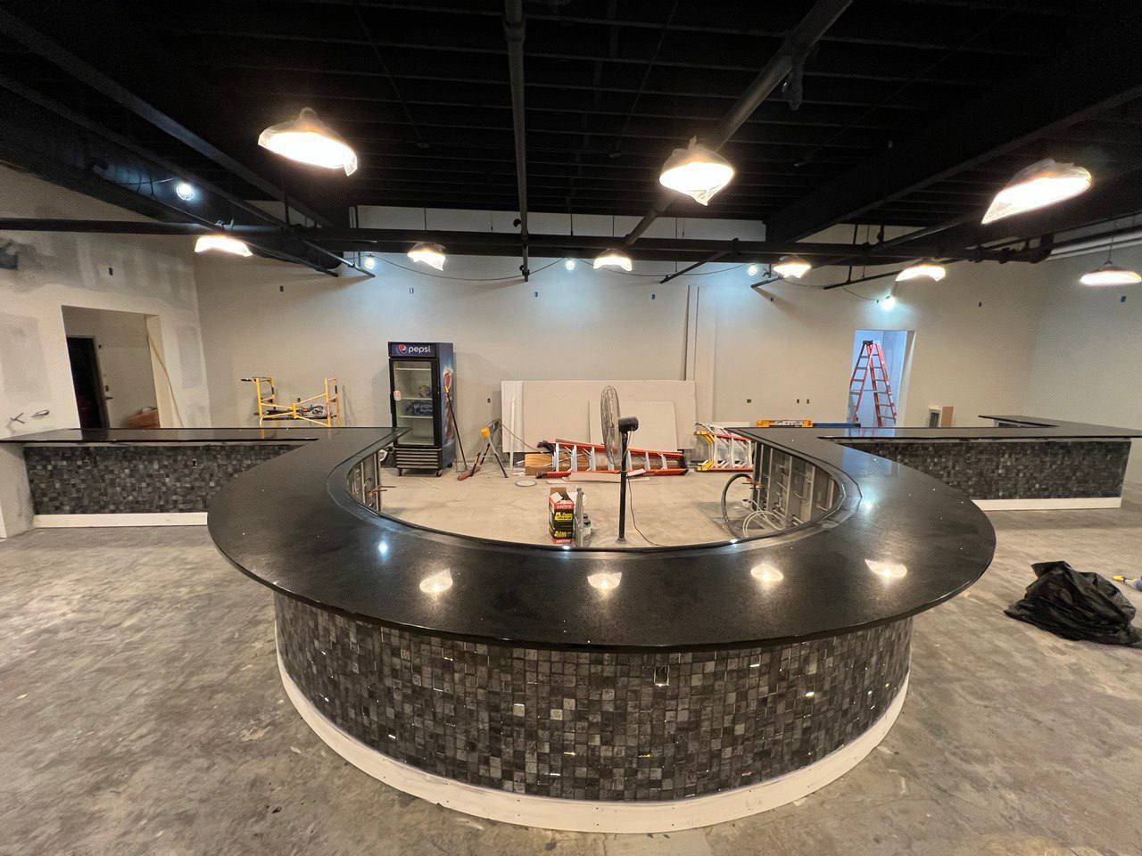 Interior of a partially constructed bar area with a large curved countertop, pendant lights above, and a tiled bar front.