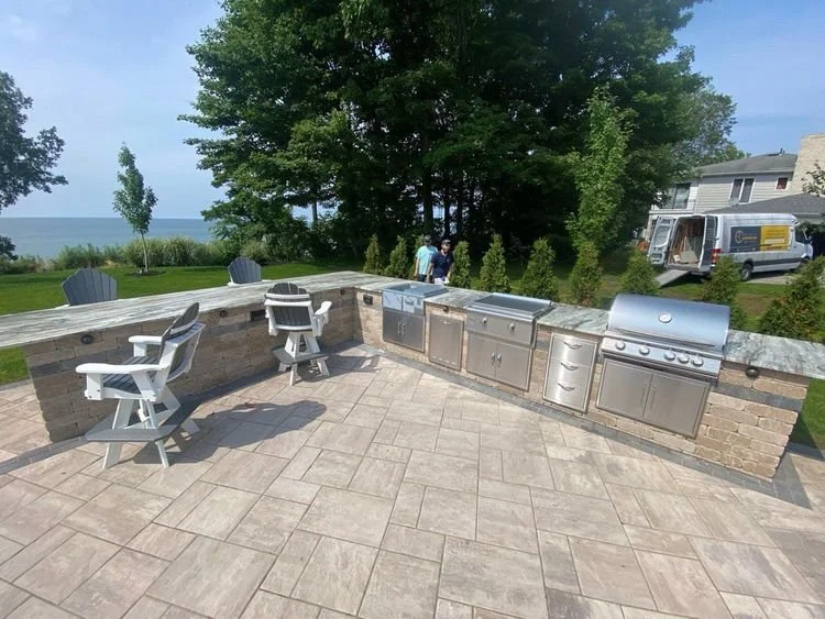 Outdoor kitchen area with stainless steel grills, granite countertops, and swivel chairs on a tiled patio, adjacent to a grassy lawn with trees.