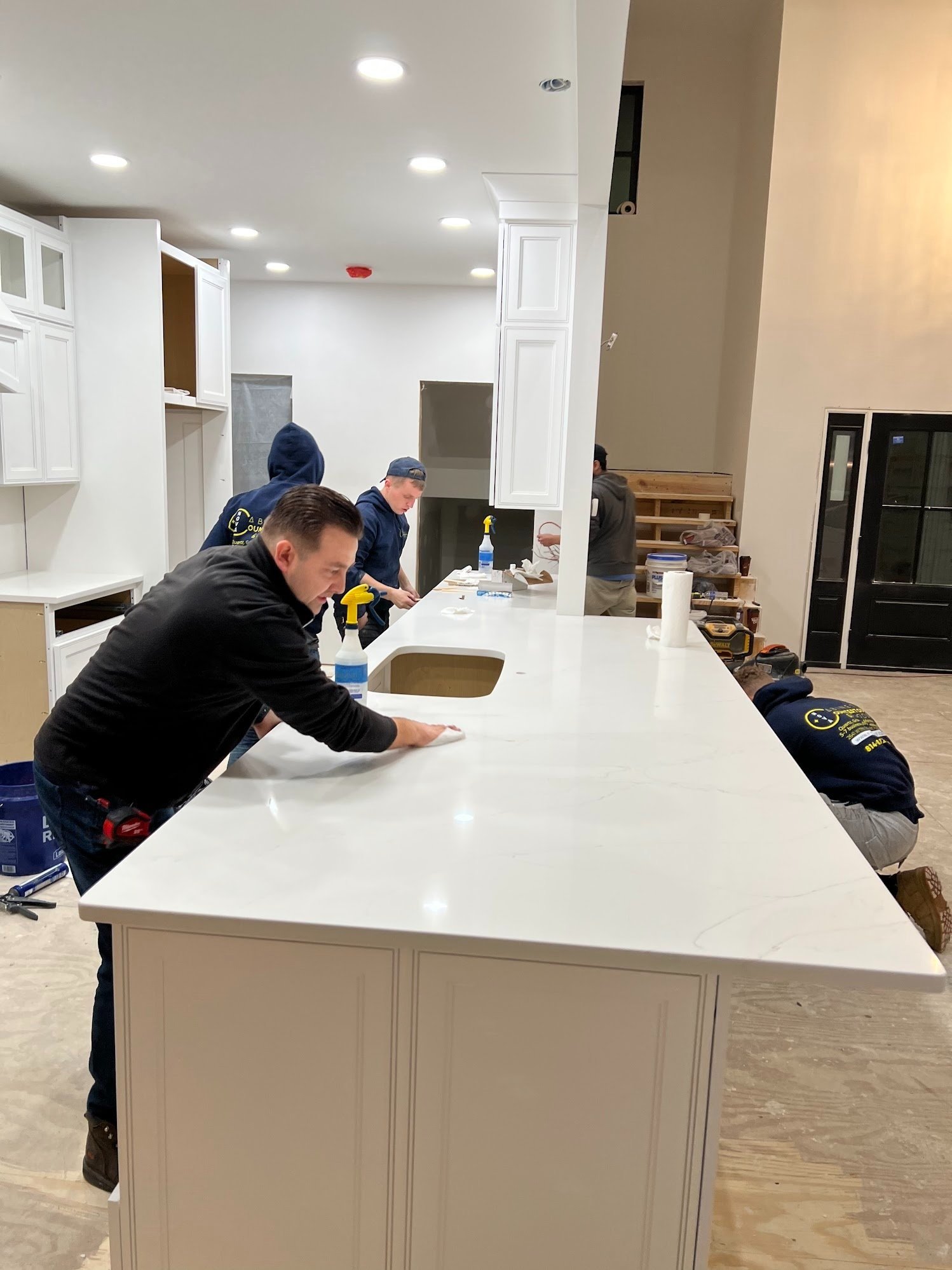 People installing a kitchen island countertop with white cabinets and recessed lighting.