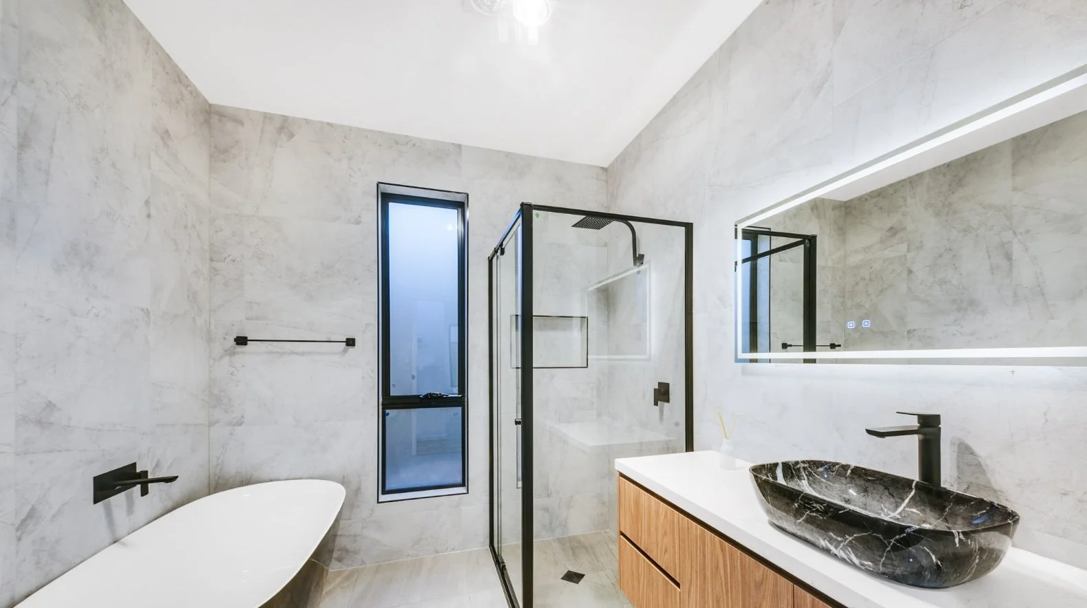 Modern bathroom with marble walls, a black marble sink, a large mirror with LED lighting, a glass shower enclosure, a window, and a freestanding bathtub.