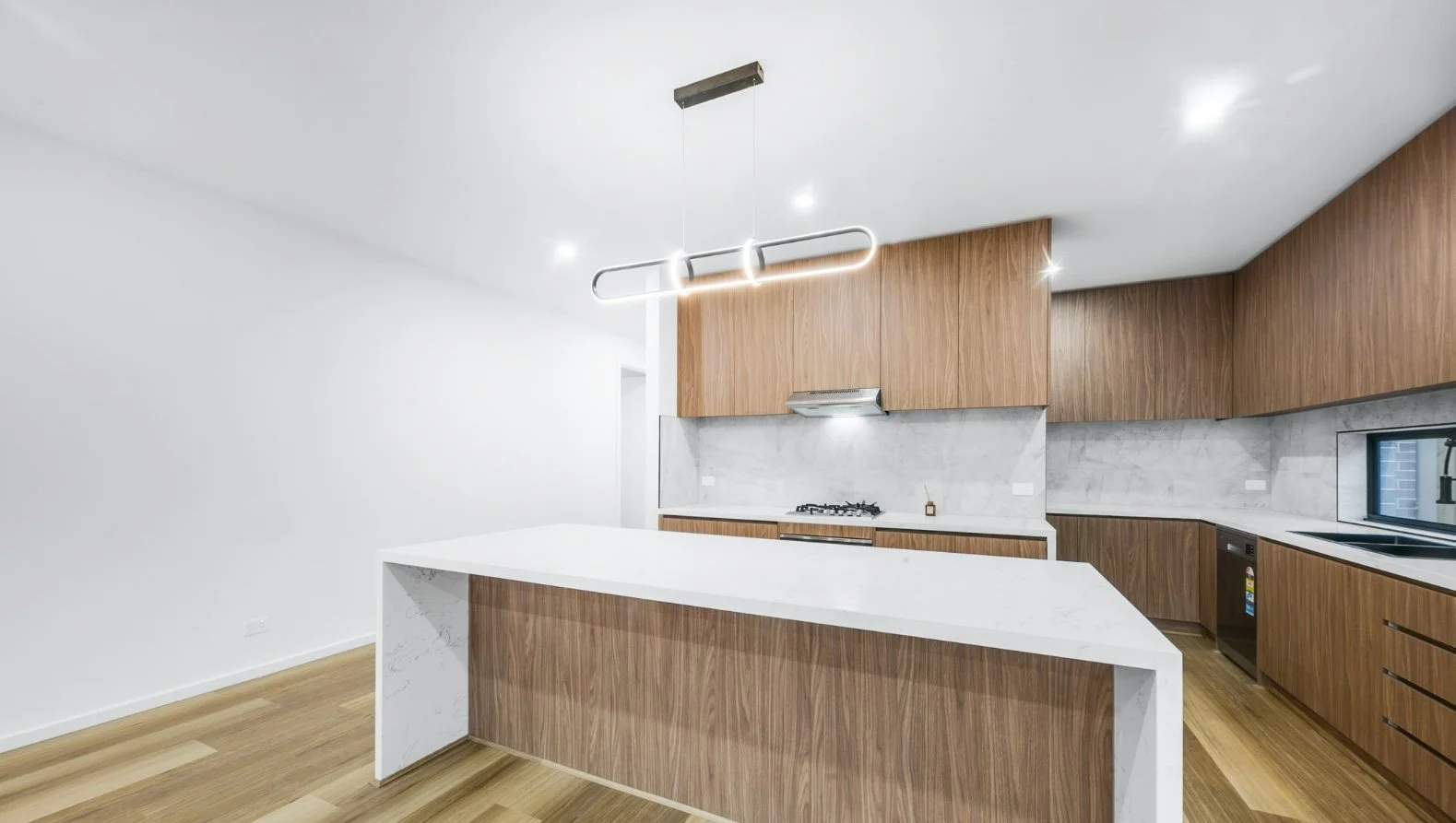 Modern kitchen with wooden cabinets, marble countertops, and a white island with marble sides. Includes a stove, sink, and dishwasher, with a minimalist chandelier hanging from the ceiling.