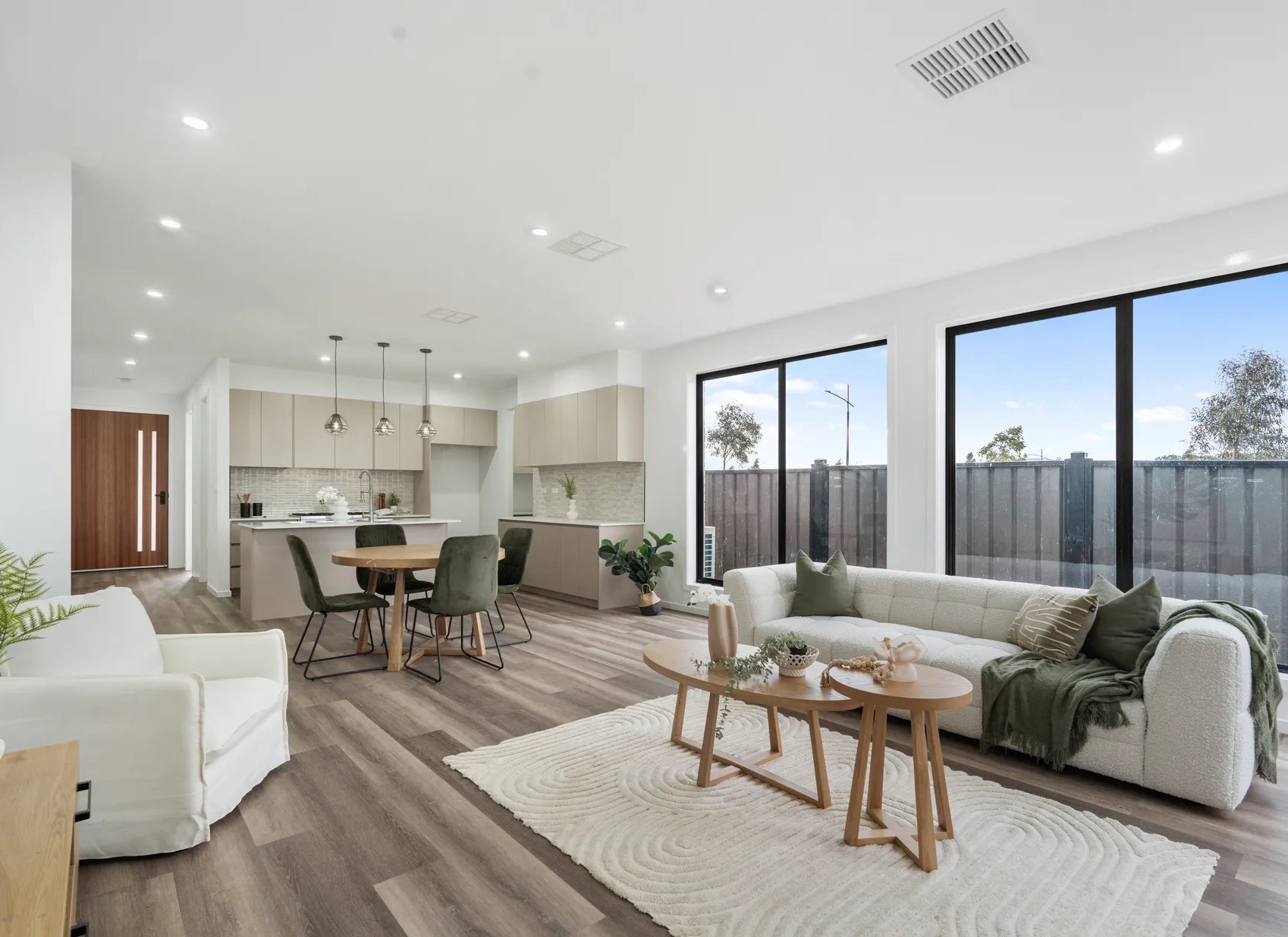 Open-concept living room and kitchen with modern decor, large windows, beige and white furniture, and wooden accents.