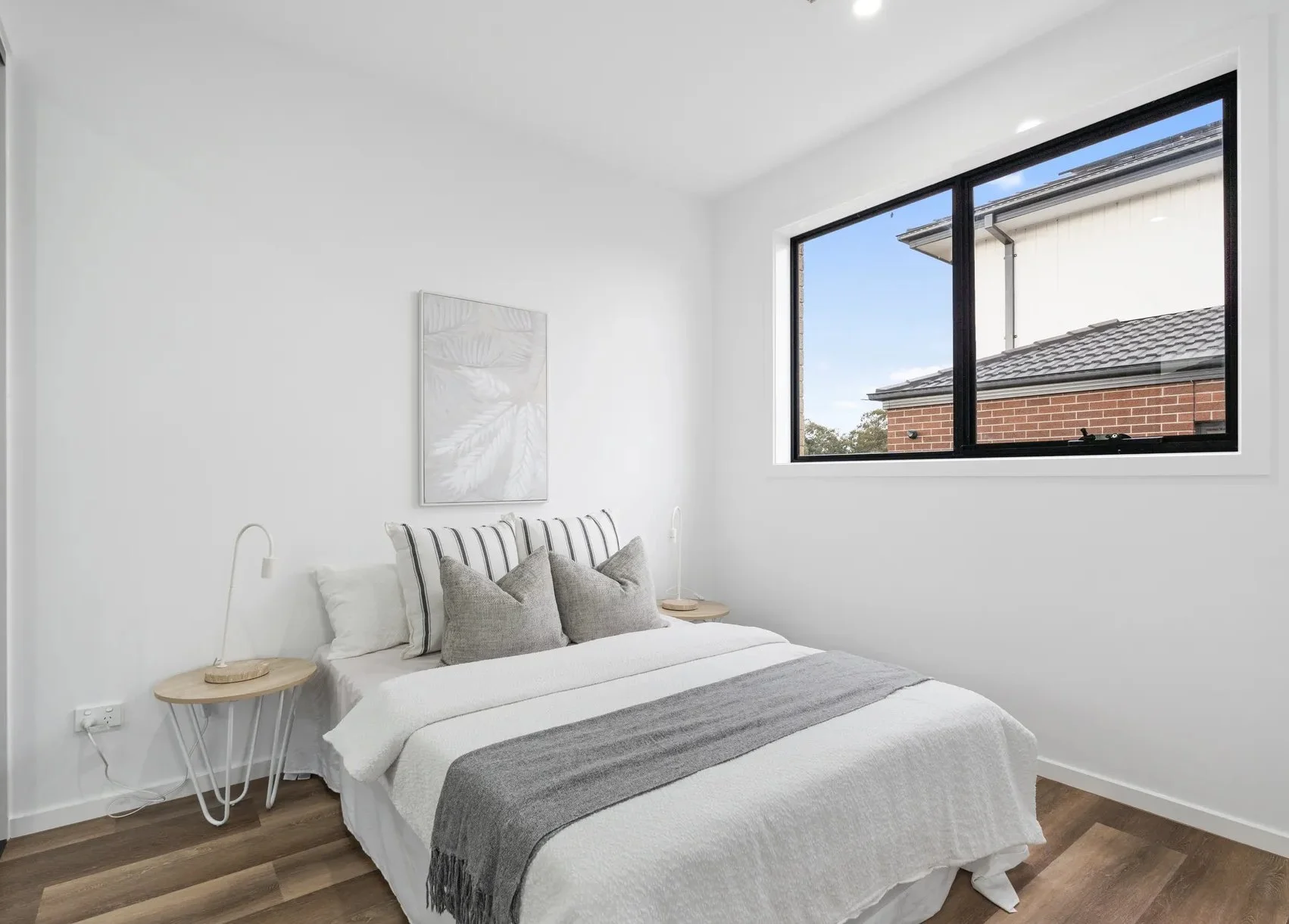 Minimalist bedroom with a white bed, gray and white throw pillows, a gray and white blanket, two round bedside tables, and a window showing a brick building outside.
