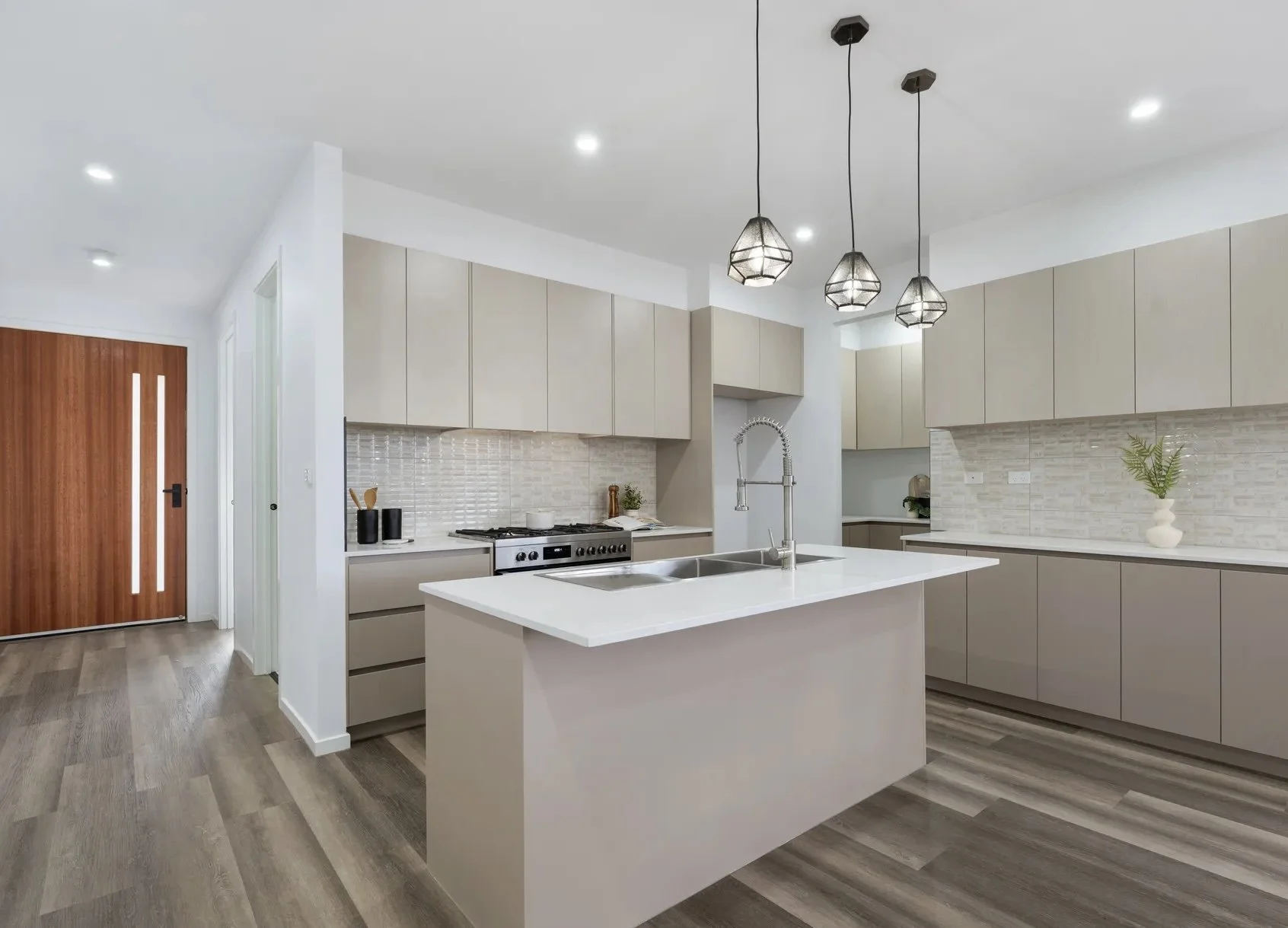 Modern kitchen with light beige cabinets, white countertops, and a central island with a sink. Pendant lights hang above the island. Wooden flooring and a front door with vertical glass panels are visible.
