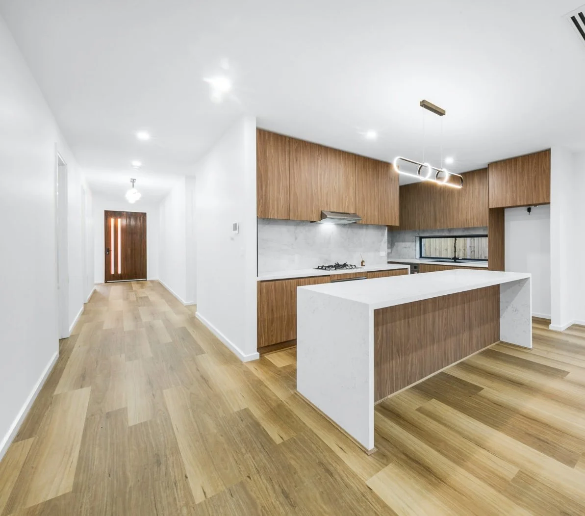 Open-concept kitchen with white and wood cabinets, marble island, wooden flooring, modern lighting, and a hallway leading to a front door.