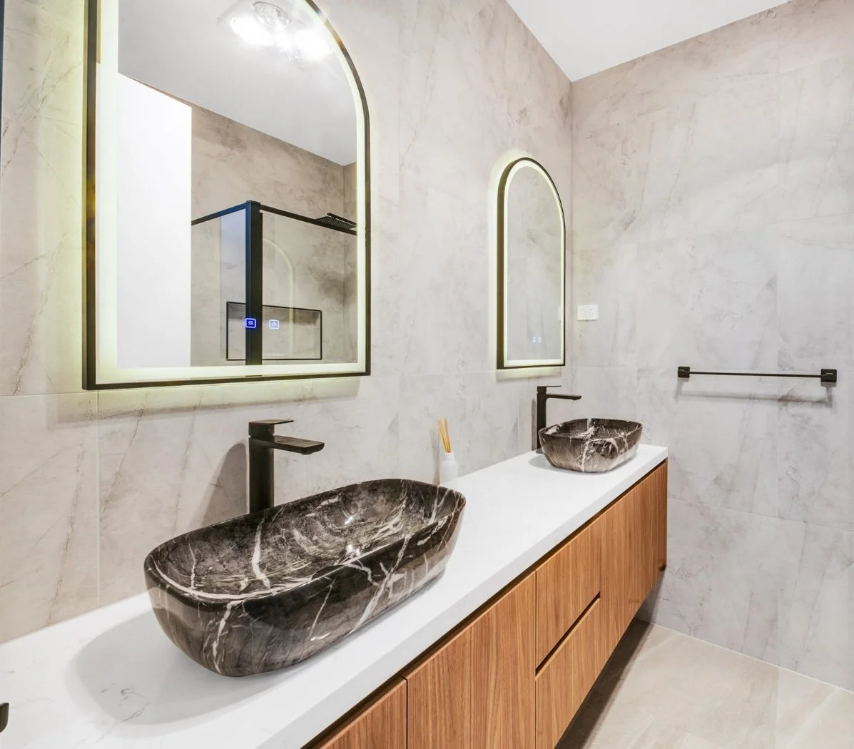 Modern bathroom with two black marble vessel sinks, wooden vanity, black faucets, illuminated arched mirrors, and beige tiled walls.