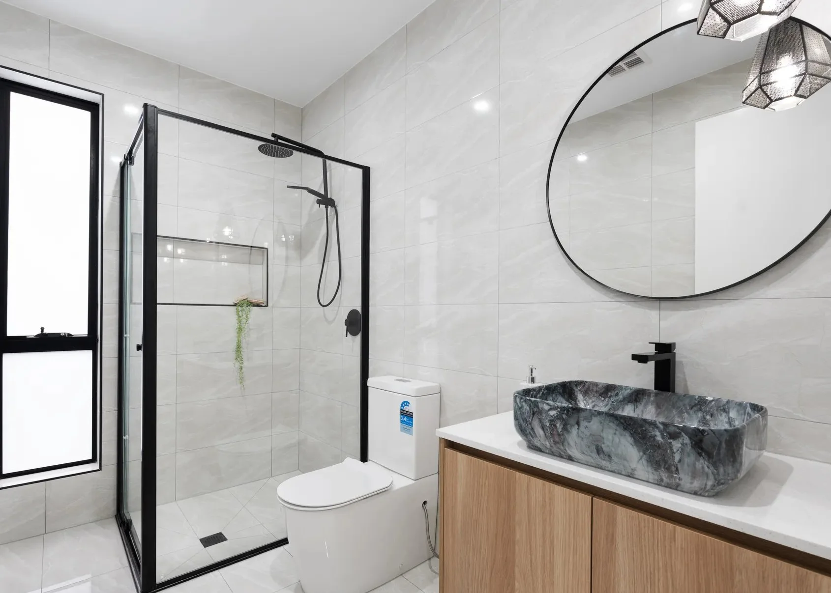Modern bathroom with a glass-enclosed shower, a white toilet, a wooden vanity with a black rectangular sink, and a round mirror on a tiled wall.