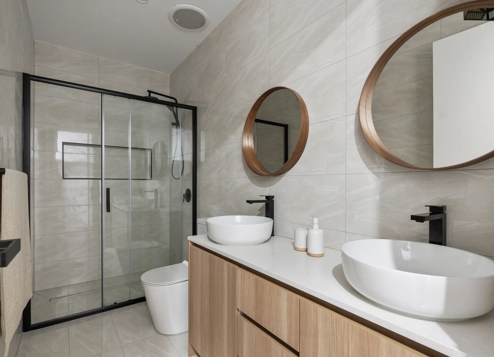 Modern bathroom with dual vessel sinks on a wooden vanity, round wall mirrors, beige tiled walls, a glass shower enclosure with black frame, and a white toilet.