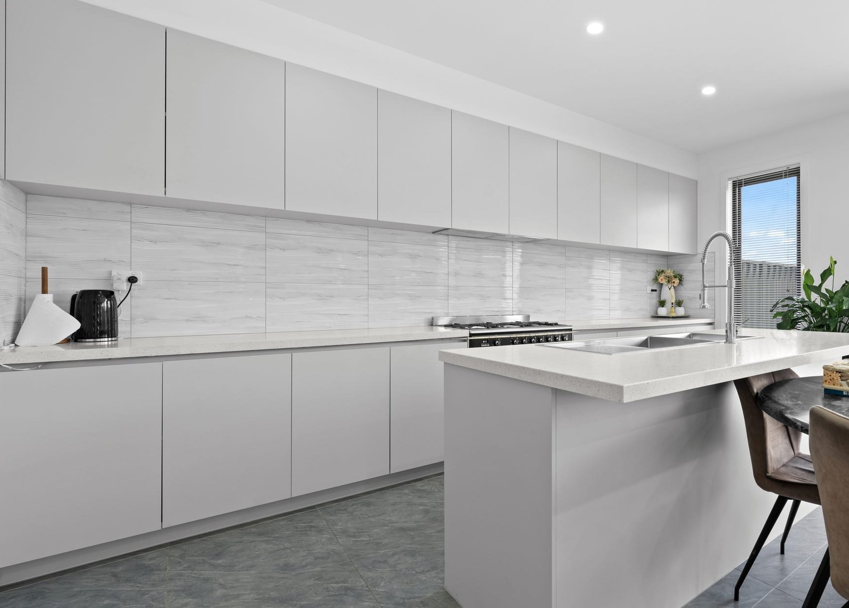 Modern white kitchen with white cabinets, marble backsplash, and a kitchen island with a double sink. There are a few plants and a small flower arrangement near the window, and some kitchen appliances on the counter.