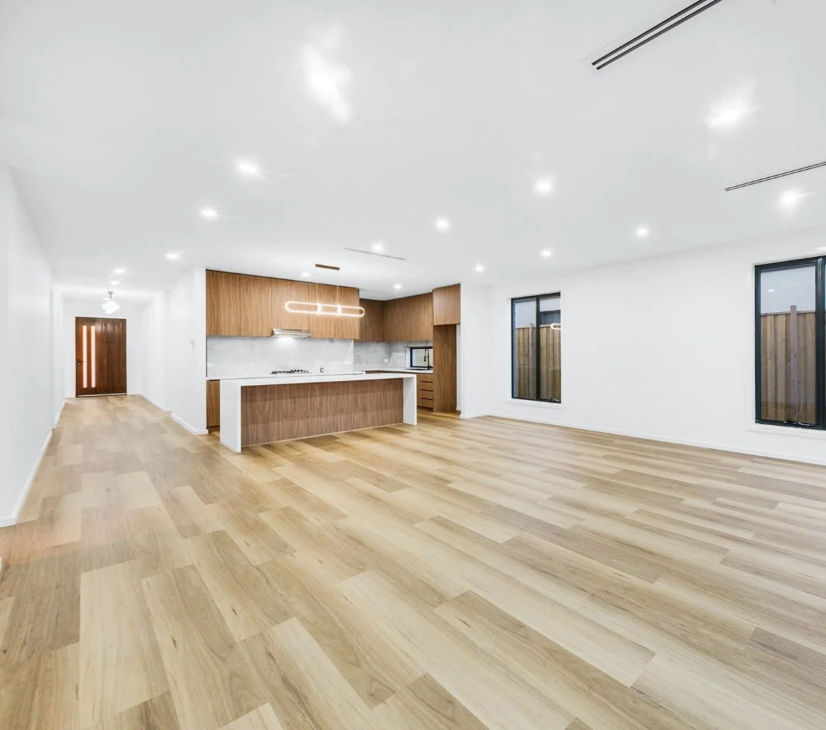 Empty modern living room with white walls, wooden flooring, and a kitchen area with wooden cabinets and a white island.