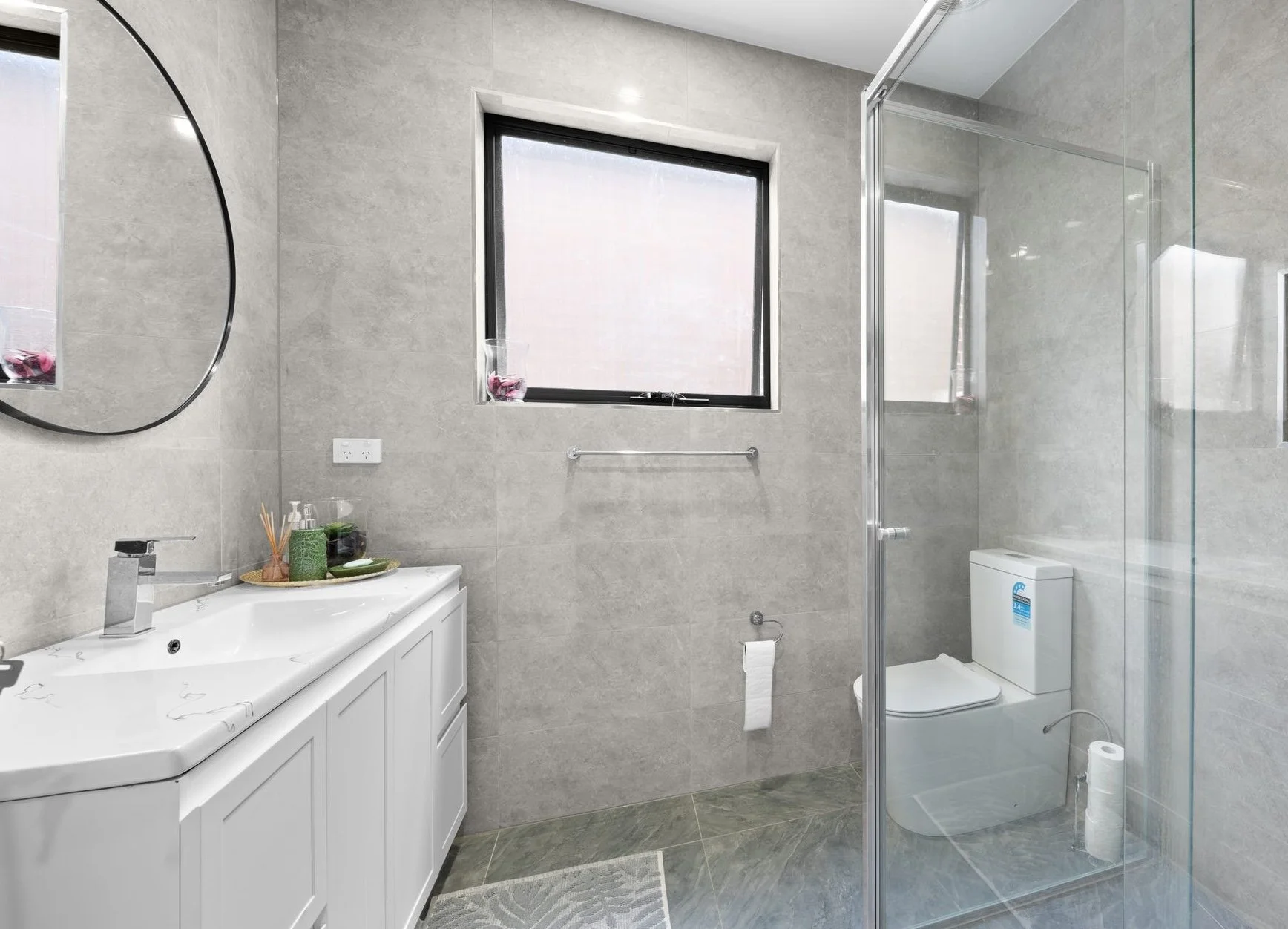 Modern bathroom with gray tiled walls and floor, a white vanity with a black faucet, a round mirror, a window with a small pink flower arrangement, a toilet inside a glass shower enclosure, and a towel rack with a hanging toilet paper.