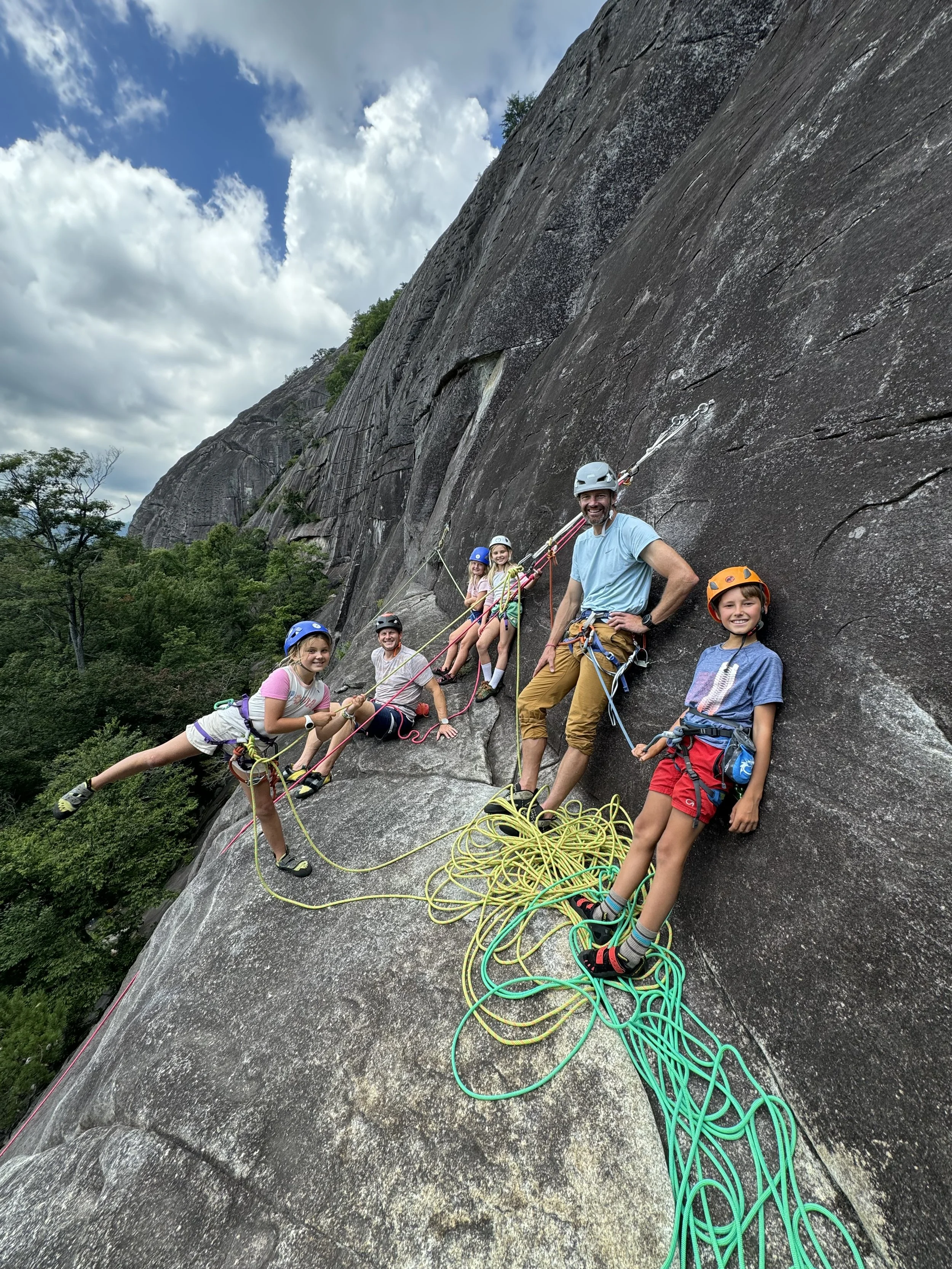 A guide and four kids smile on the granite face of Looking Glass Rock during a family guided rock climbing day with Pisgah Climbing School in Brevard, NC