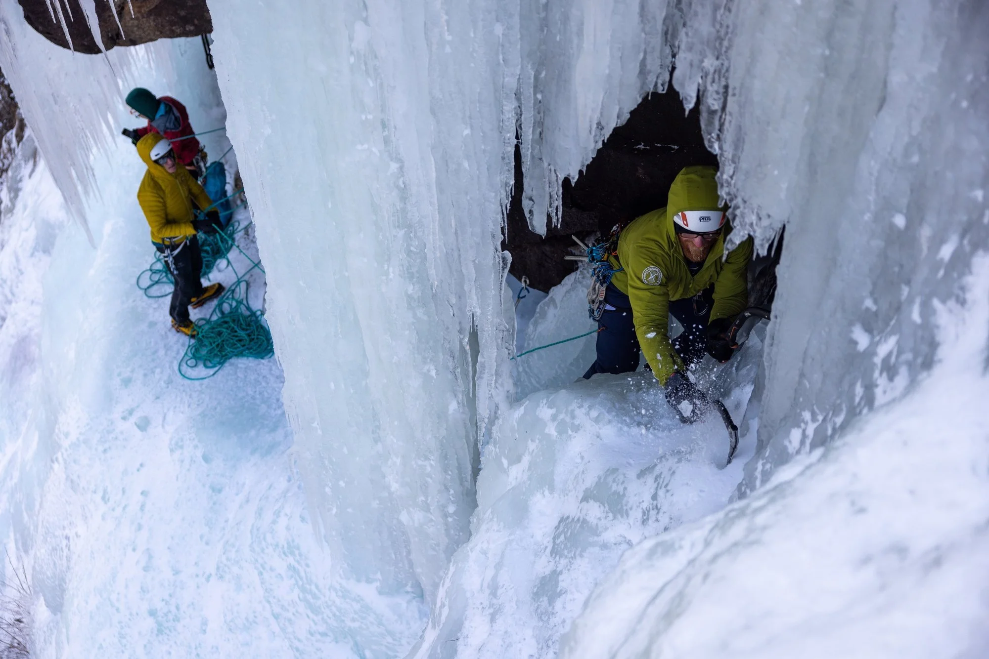 Ice climber swinging tools through the icicle cave on Standard Route, Frankenstein Cliff, New Hampshire, with partners belaying below