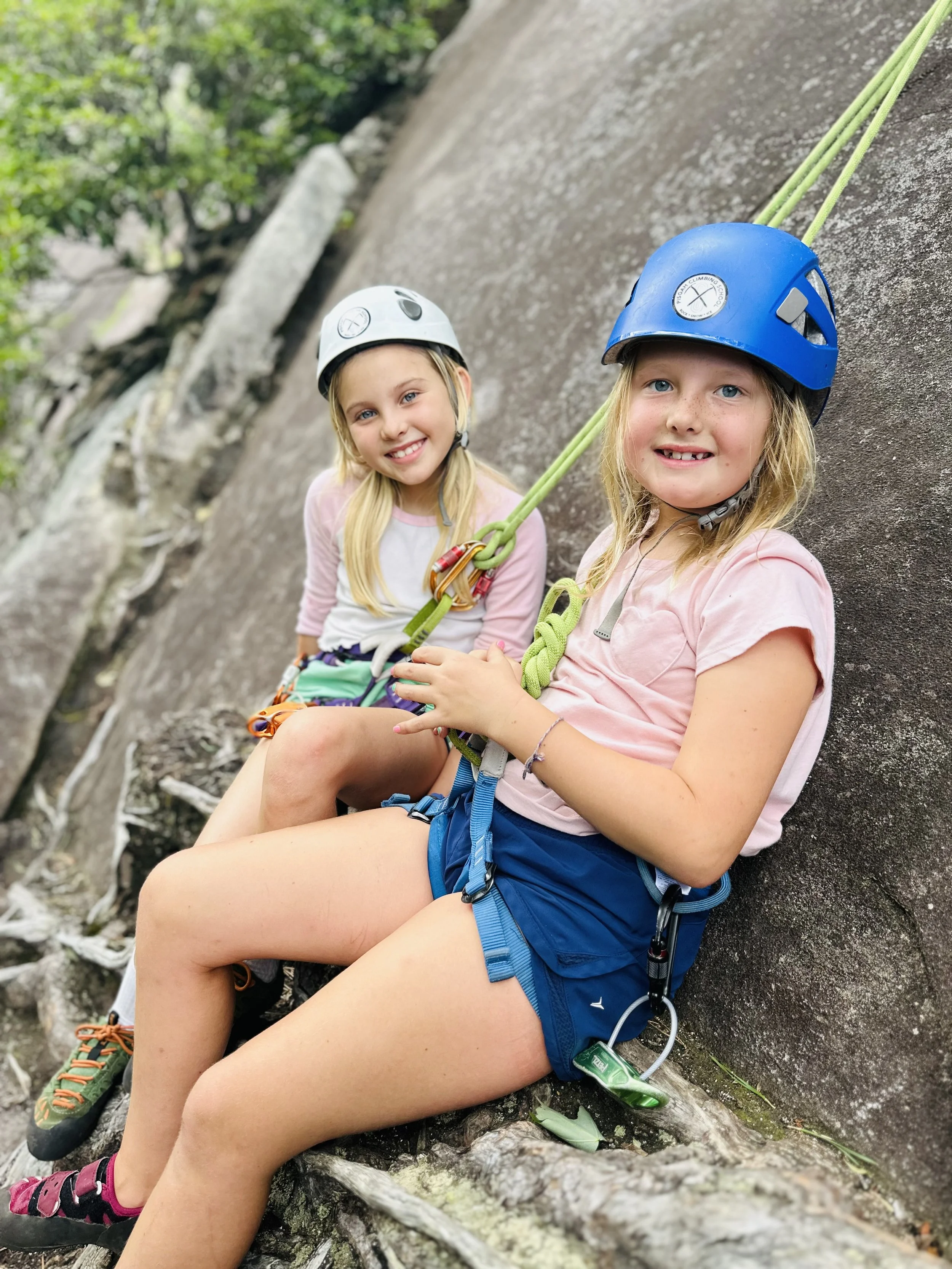 Two young girls in helmets and harnesses smiling on the granite rock face during a kids guided rock climbing lesson with Pisgah Climbing School in Pisgah National Forest, NC