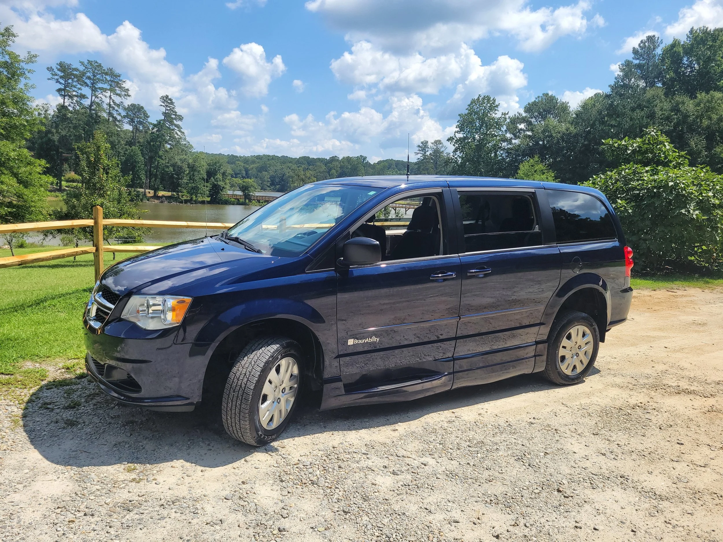 Black minivan parked outdoors near a grassy area with a wooden fence, a river, and green trees in the background. The sky is partly cloudy with scattered white clouds.