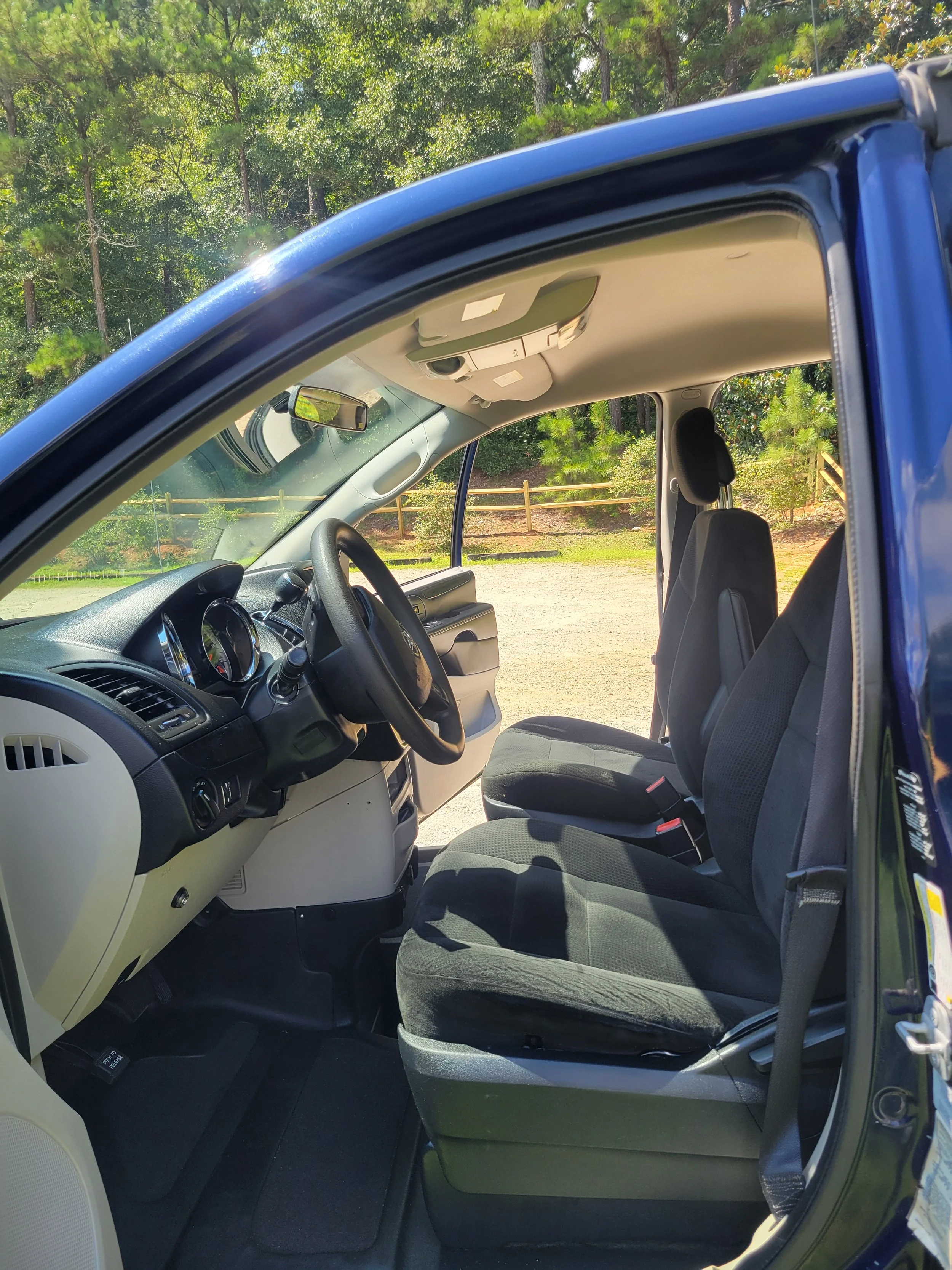 Inside view of a blue vehicle showing the driver's seat, passenger seats, steering wheel, and dashboard, with a background of green trees.