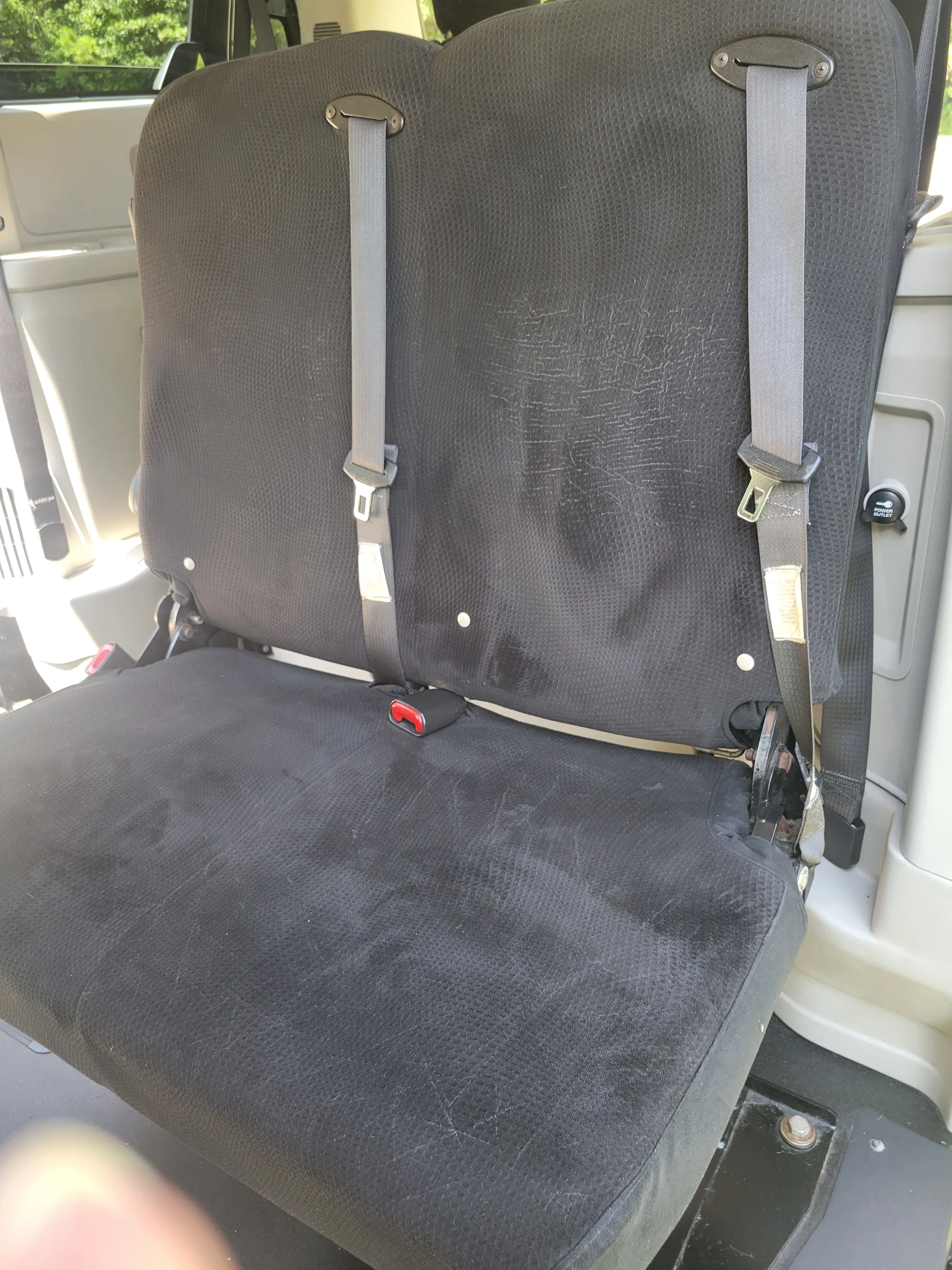 The back seat of a vehicle with a black fabric seat cover and two seat belts, with part of the vehicle's interior and a window showing greenery outside.