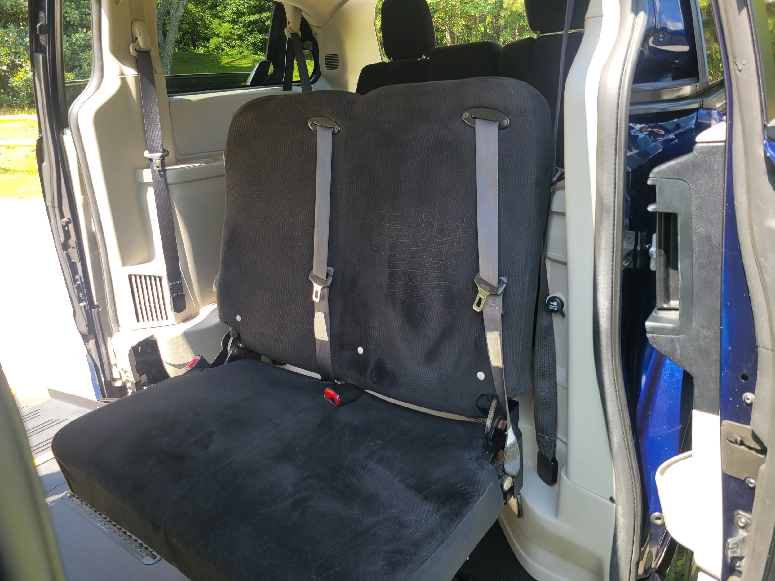 Interior view of a vehicle showing black fabric seats with gray seat belts, with a background of green trees and bright sunlight outside.