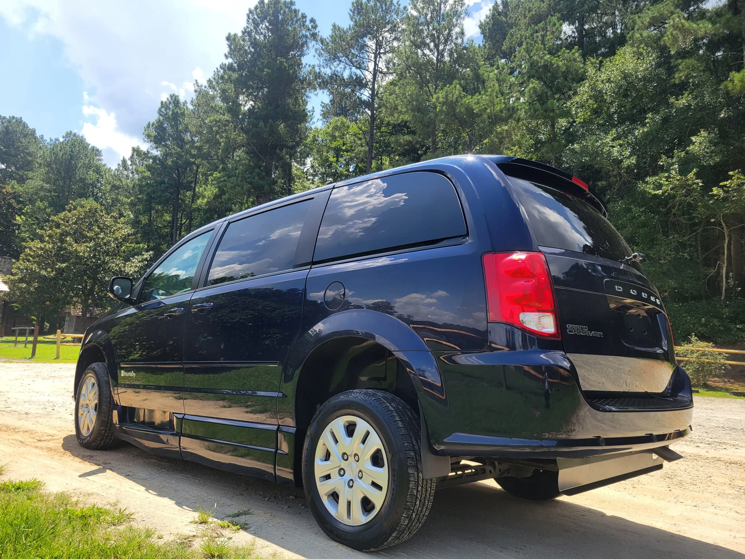 A black minivan parked on a dirt lot surrounded by trees under a blue sky with some clouds.