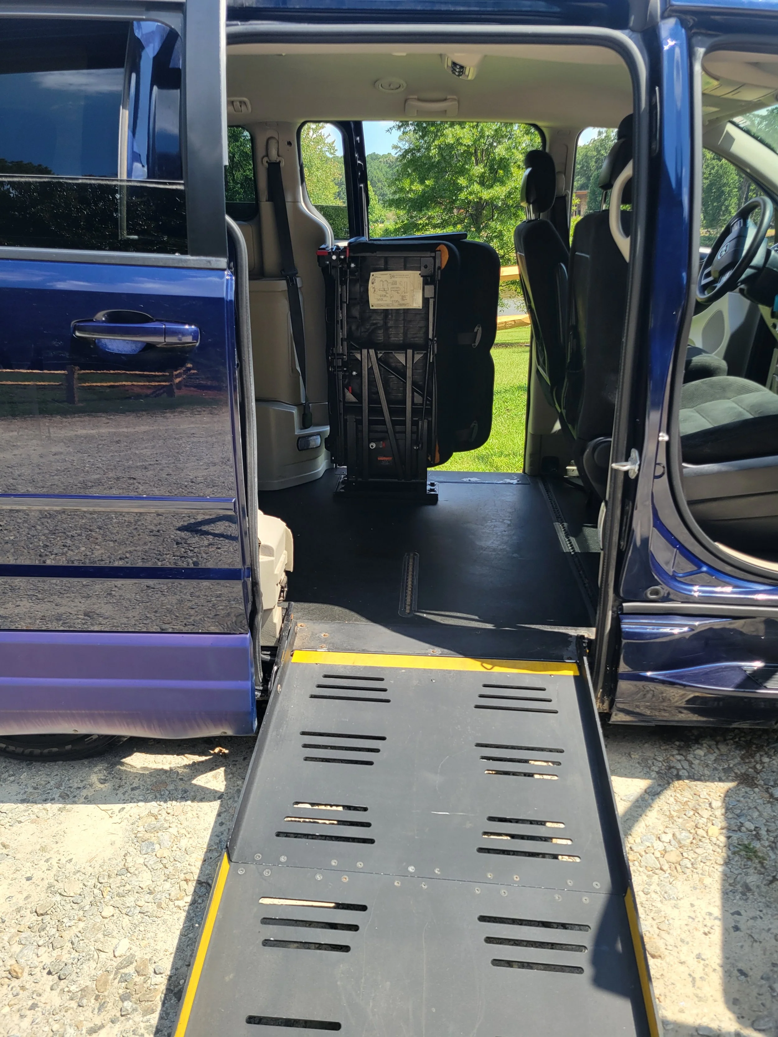 A blue van with its sliding door open, revealing an interior wheelchair ramp extending onto the ground with a gravel surface. The van has two front seats and large windows showing a green outdoor landscape with trees.