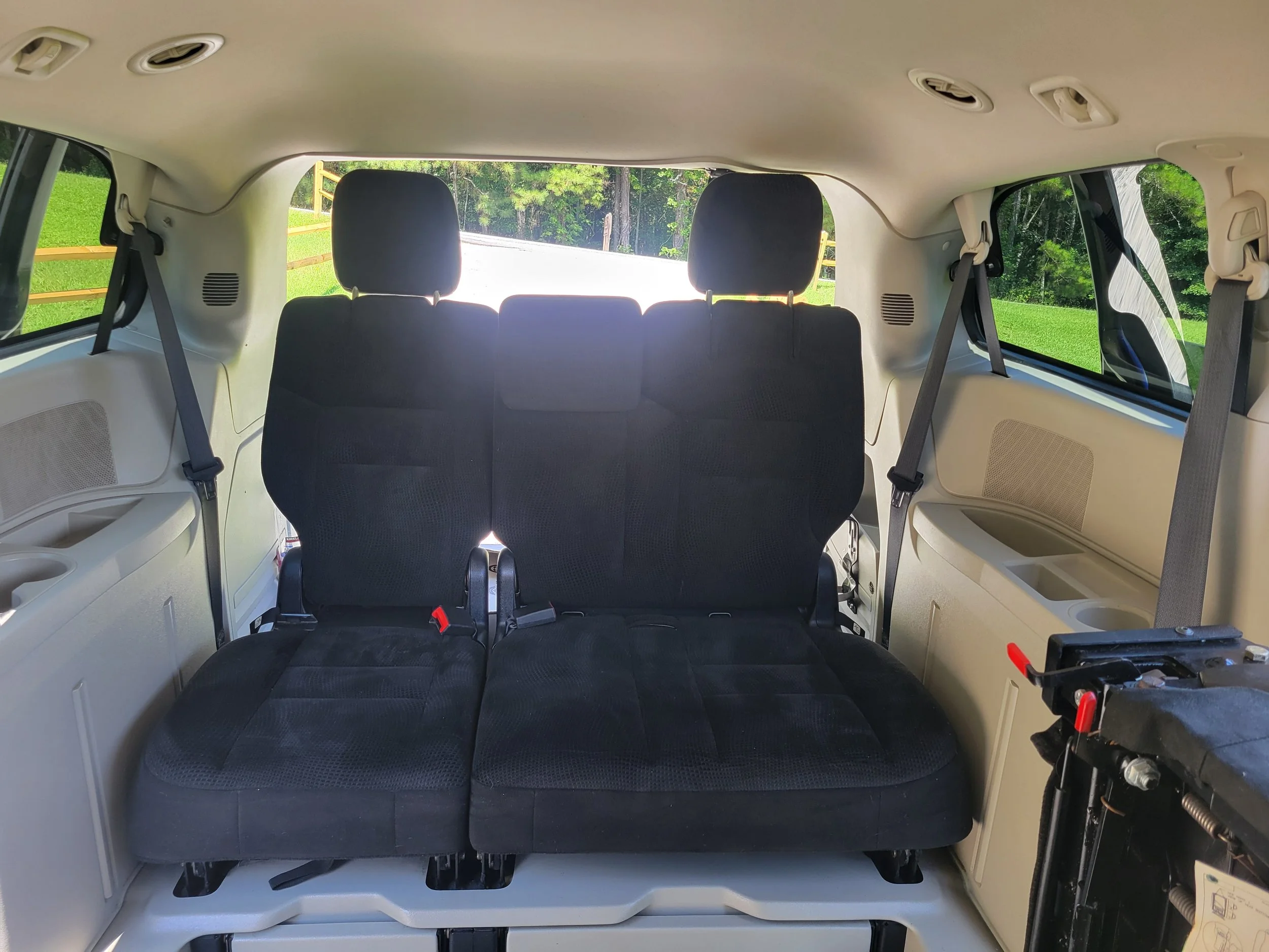 Interior view of a vehicle's rear with black fabric seats, side windows showing green grass and trees outside, and various interior fixtures, including seat belts and vents.