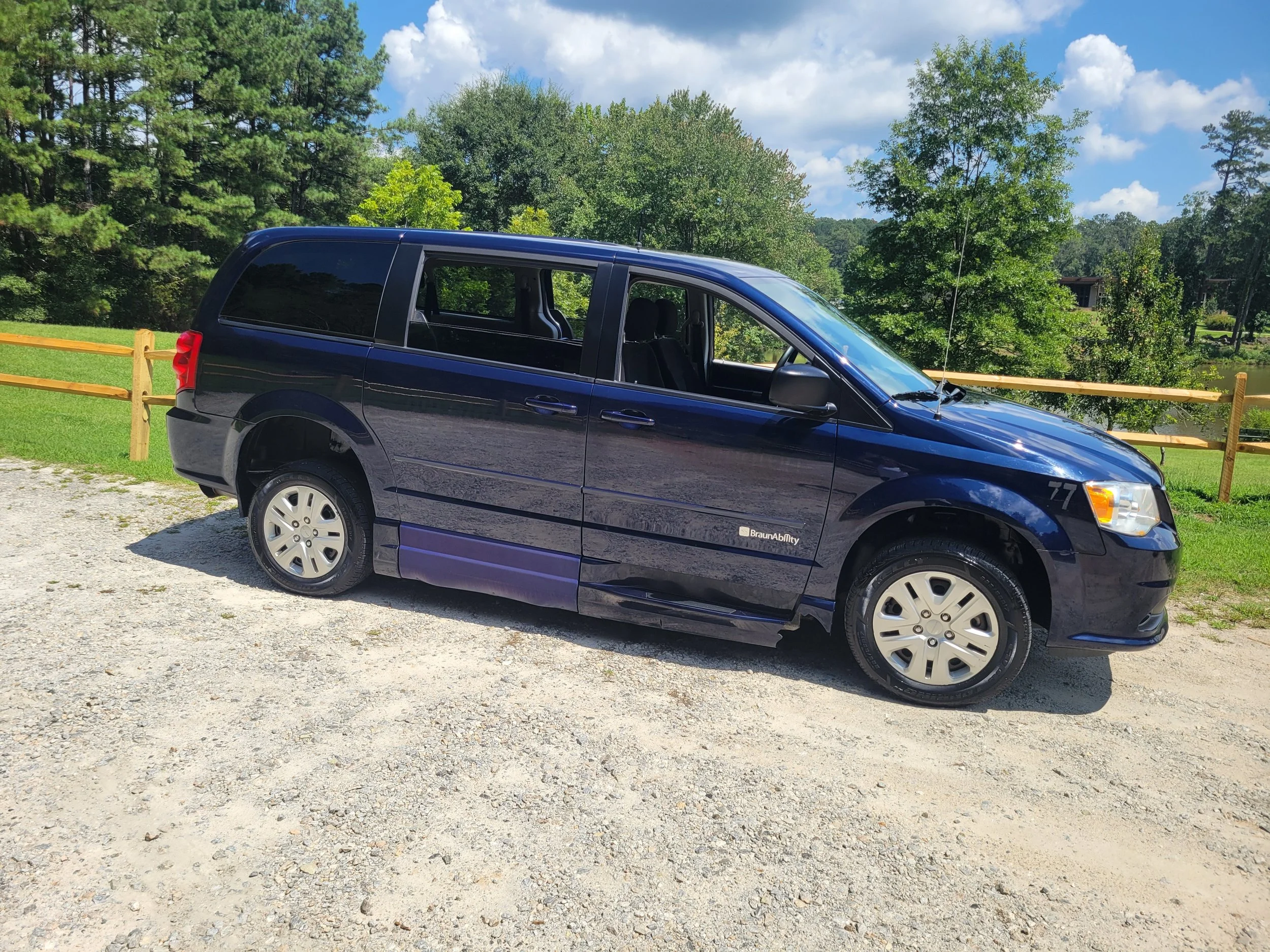 A dark blue minivan parked on a gravel area near a wooden fence, with trees, a pond, and a house in the background under a partly cloudy sky.