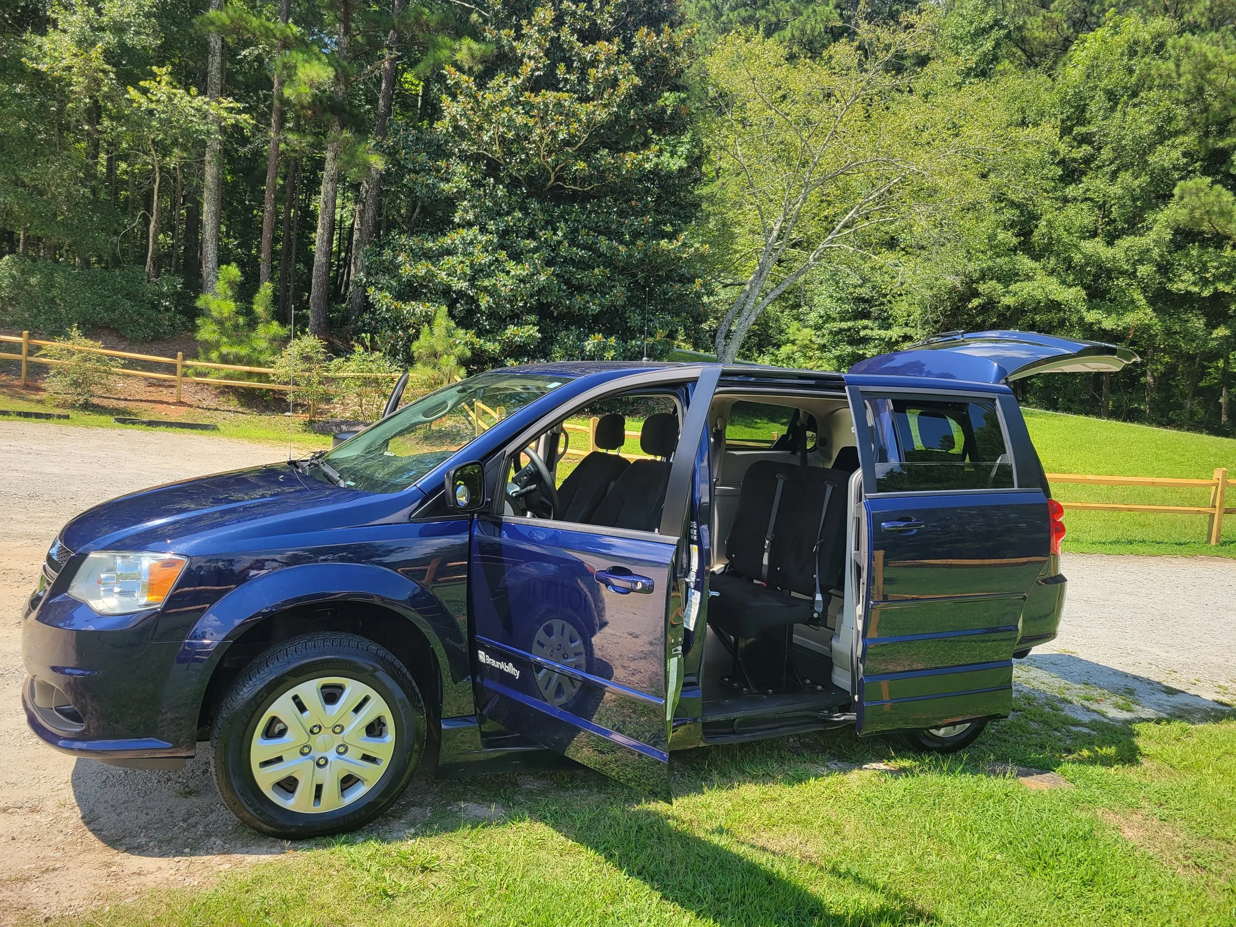 A blue minivan with its side sliding door open, parked on a grassy area next to a gravel pathway, with a backdrop of dense green trees.