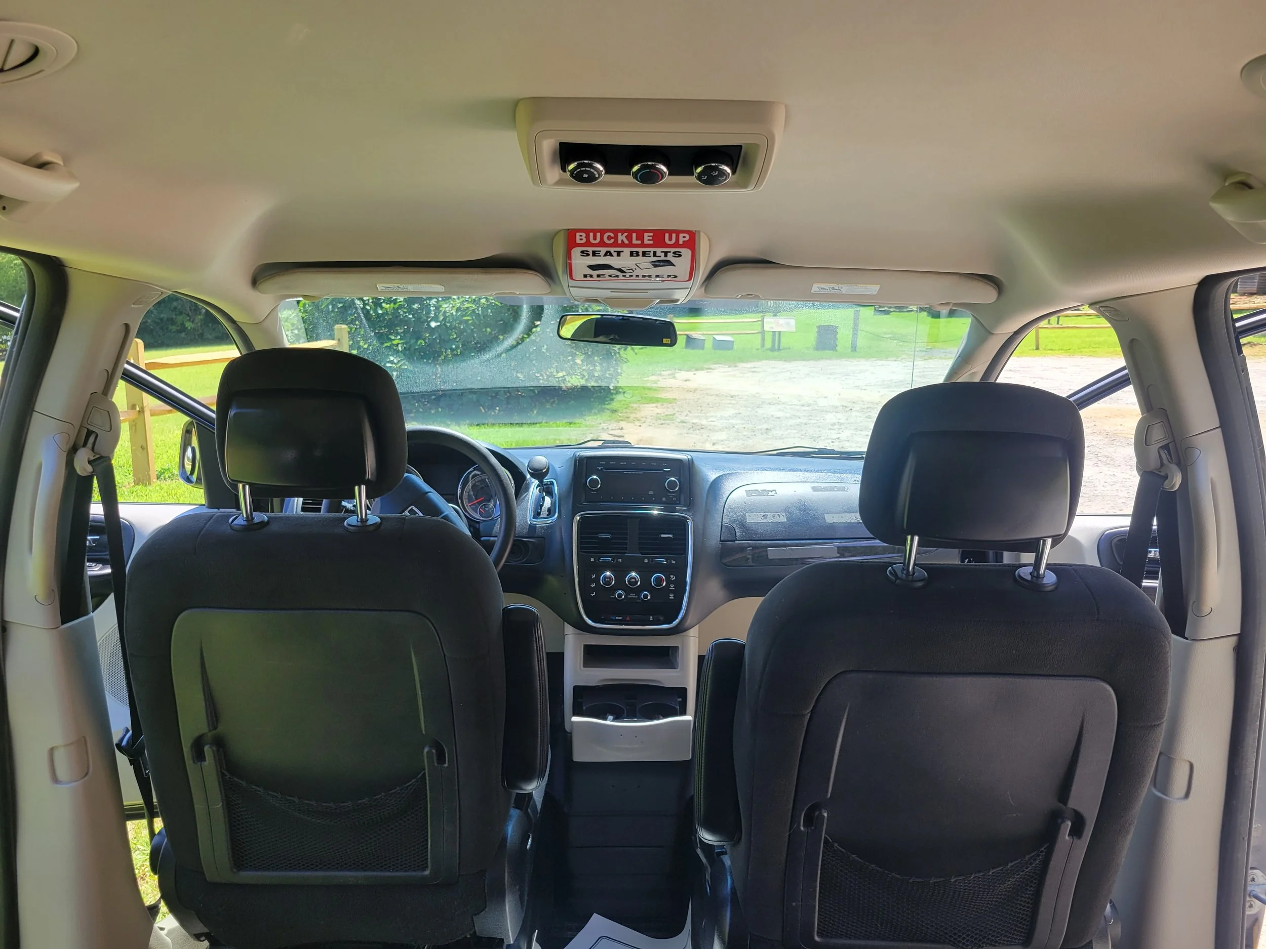 Interior of a vehicle looking forward, showing the back of front seats, dashboard, and windshield. Outside, there is a grassy area with a yellow fence.