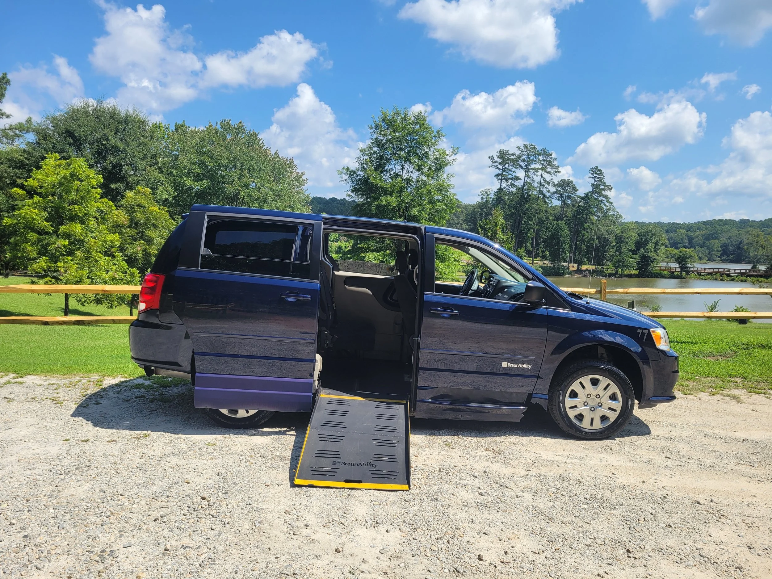 A blue van with its sliding door open and a ramp extended onto a gravel parking area, with trees and a river in the background under a partly cloudy sky.