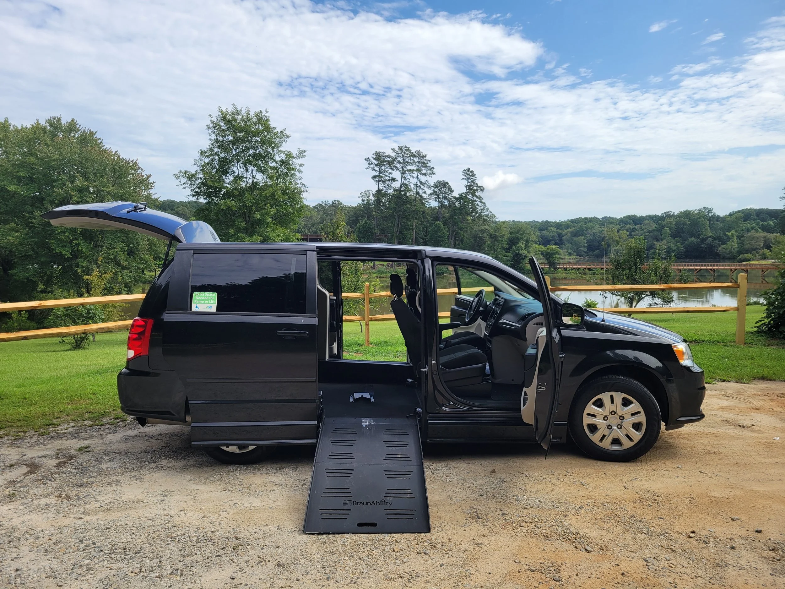 A black minivan with one sliding door open, parked on a gravel area near a body of water. There is a sign on the window indicating extra space needed for a ramp or lift. The background features green trees, a lake, and a blue sky with some clouds.
